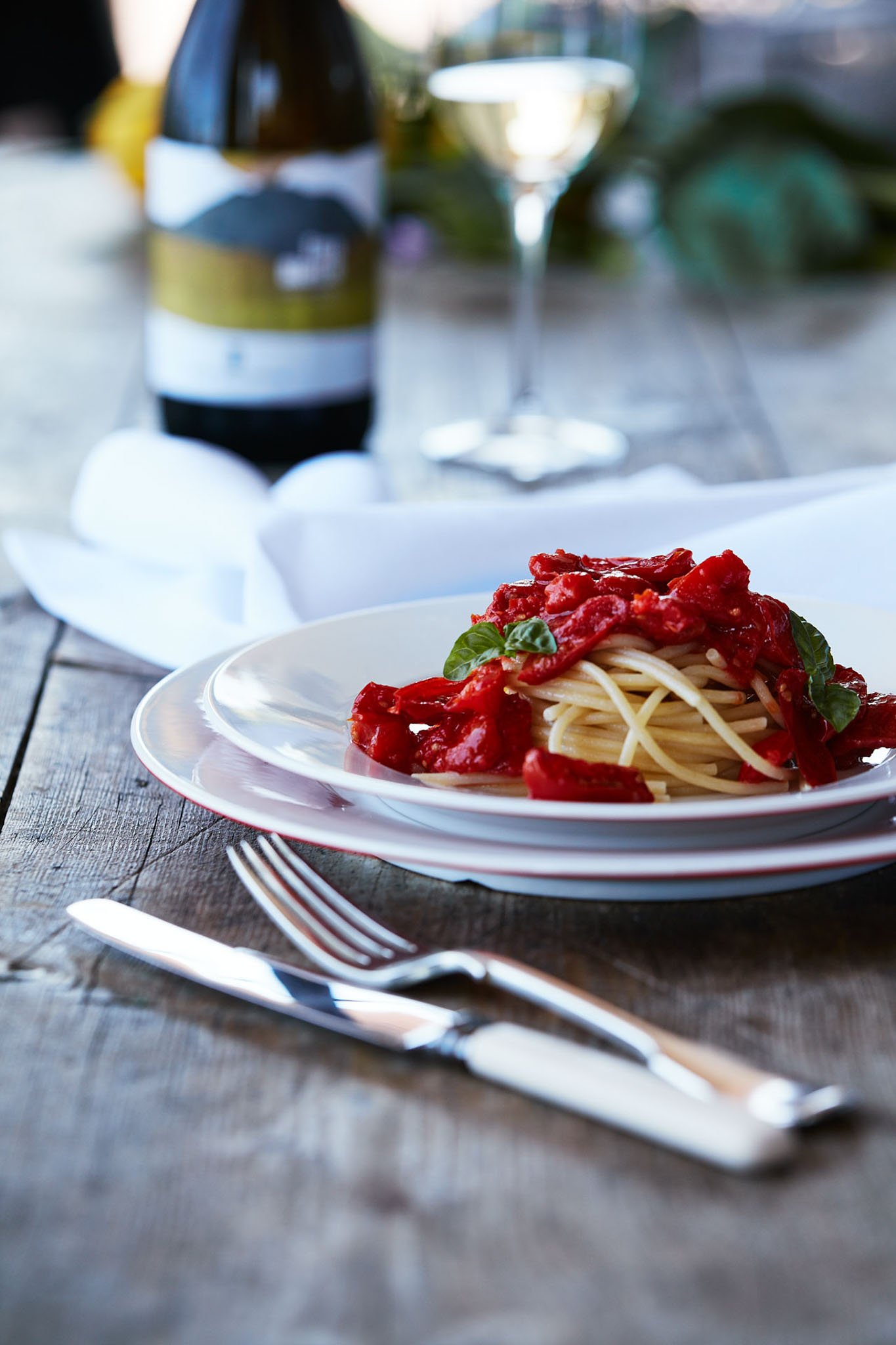 Plate of spaghetti with tomato sauce and basil, set on a rustic wooden table with a fork and knife, a glass of white wine, and a bottle of wine in the background.