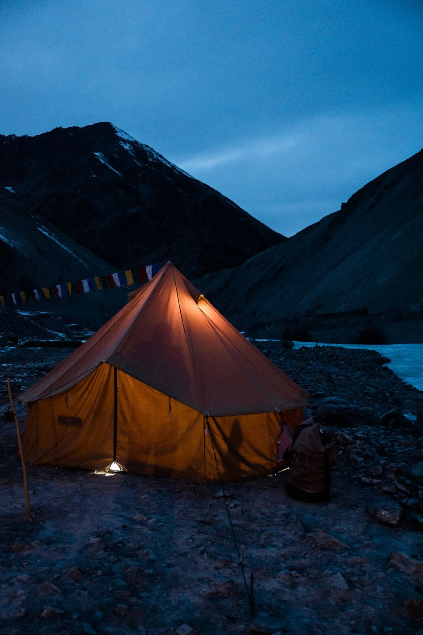 A tent illuminated from inside is set up on rocky terrain in a mountain valley at dusk. Mountains with snow patches are visible in the background, and prayer flags hang above the tent.