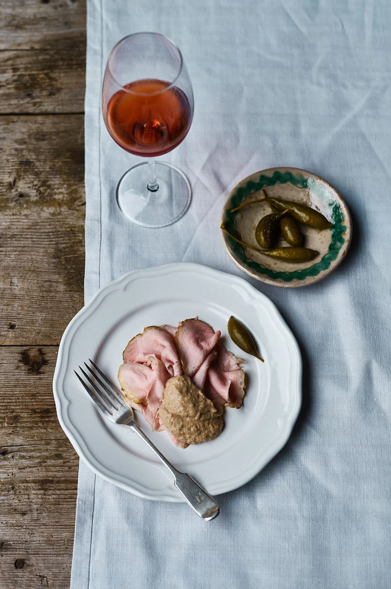 A plate with slices of ham and a dollop of mustard, a glass of rosé wine, a small bowl of pickles, and a white tablecloth on a wooden table.