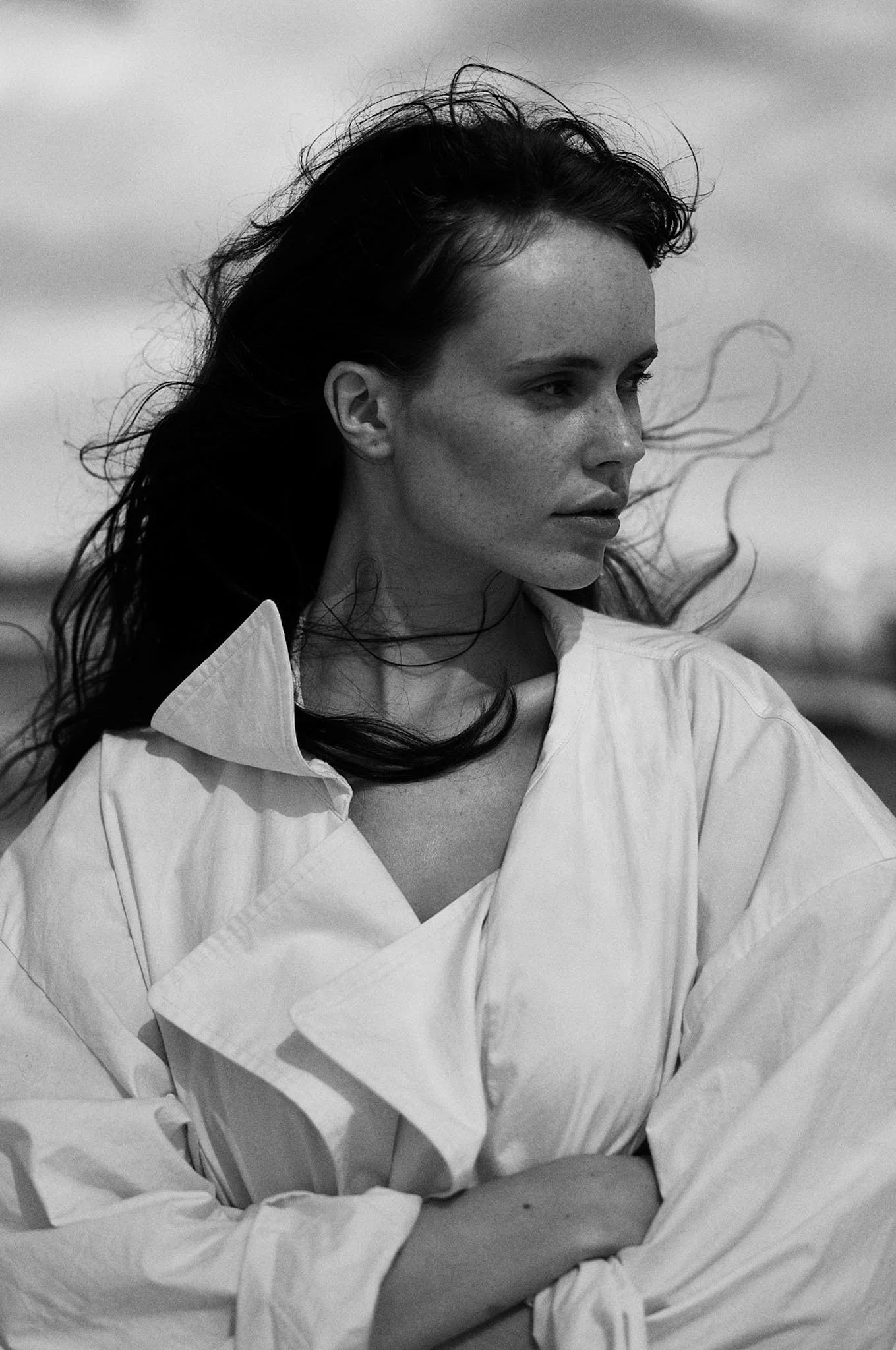 A black and white photograph of a woman with long dark hair, wearing a loose white shirt, standing outdoors with her arms crossed, looking to the side with a contemplative expression.