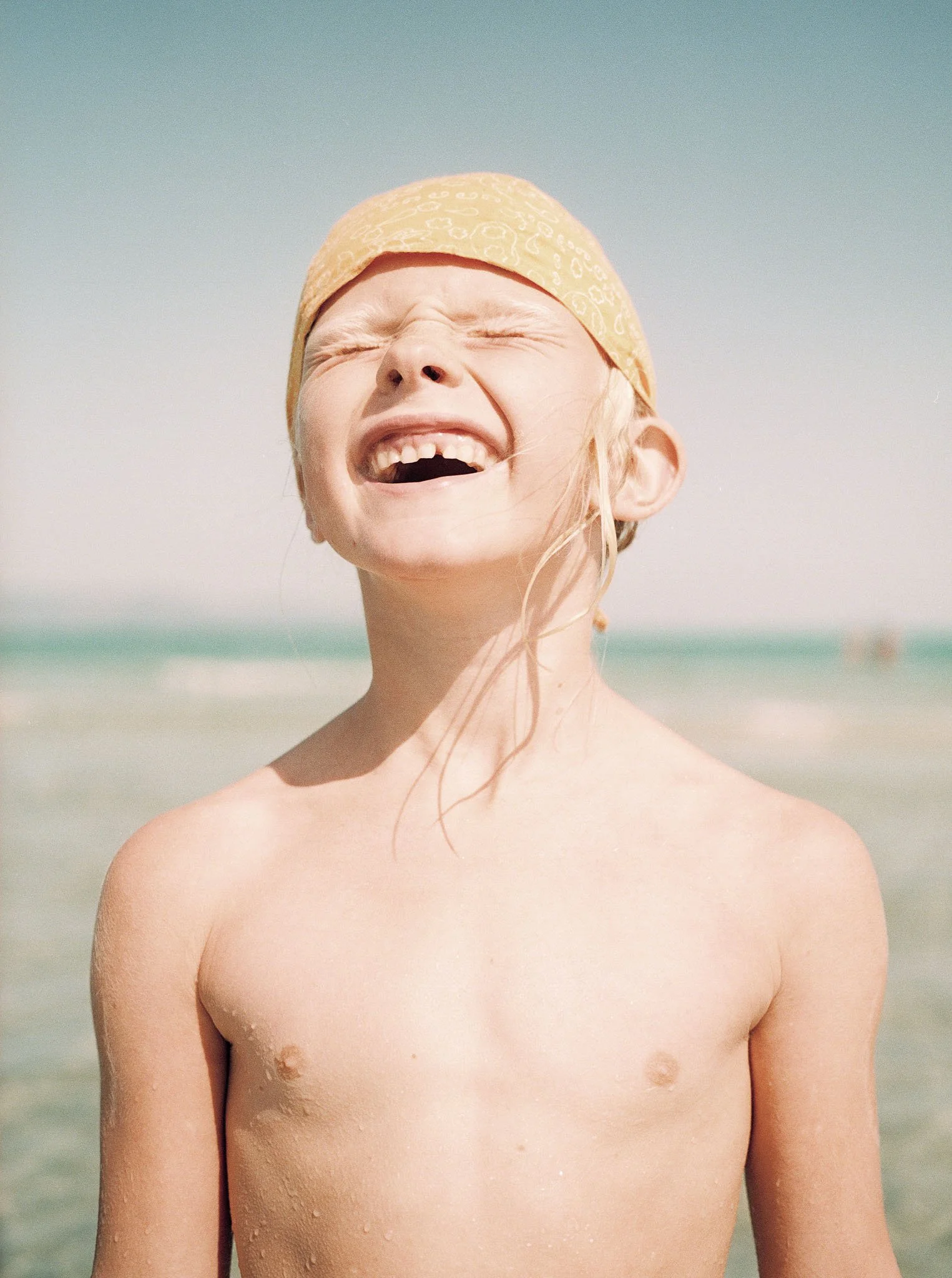 A young girl at the beach, smiling with her eyes closed, wearing a yellow bandana, and wet hair, with the ocean in the background.
