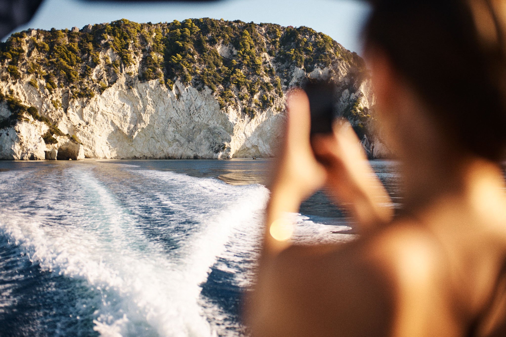 A person taking a photo with a phone while on a boat, with a view of rocky cliffs and water in the background.