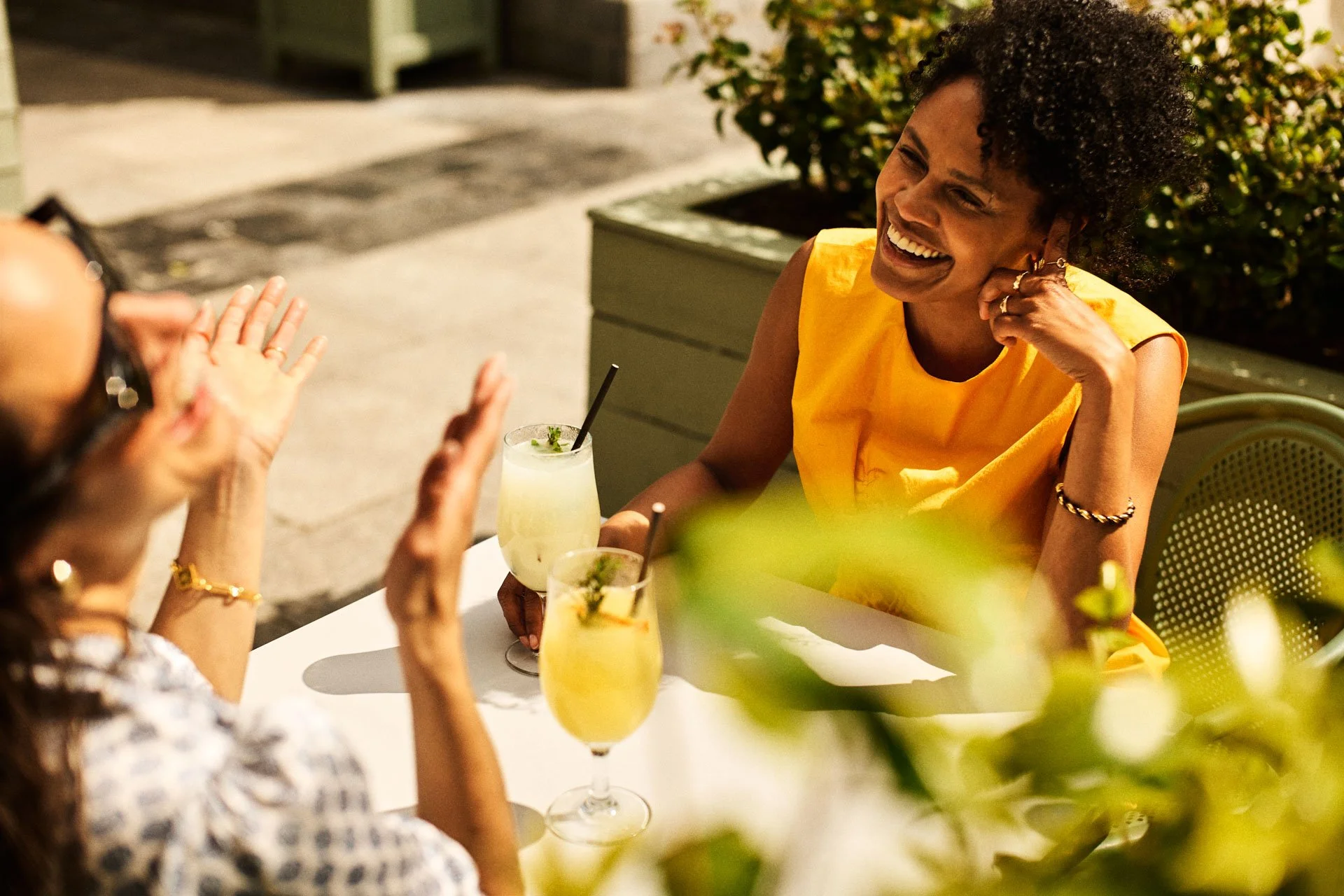 Two women are sitting at an outdoor table, engaged in conversation. One woman, wearing a bright yellow top, is smiling and laughing, while the other woman, whose face is partially visible, is gesturing with her hands. There are two glasses of a yello