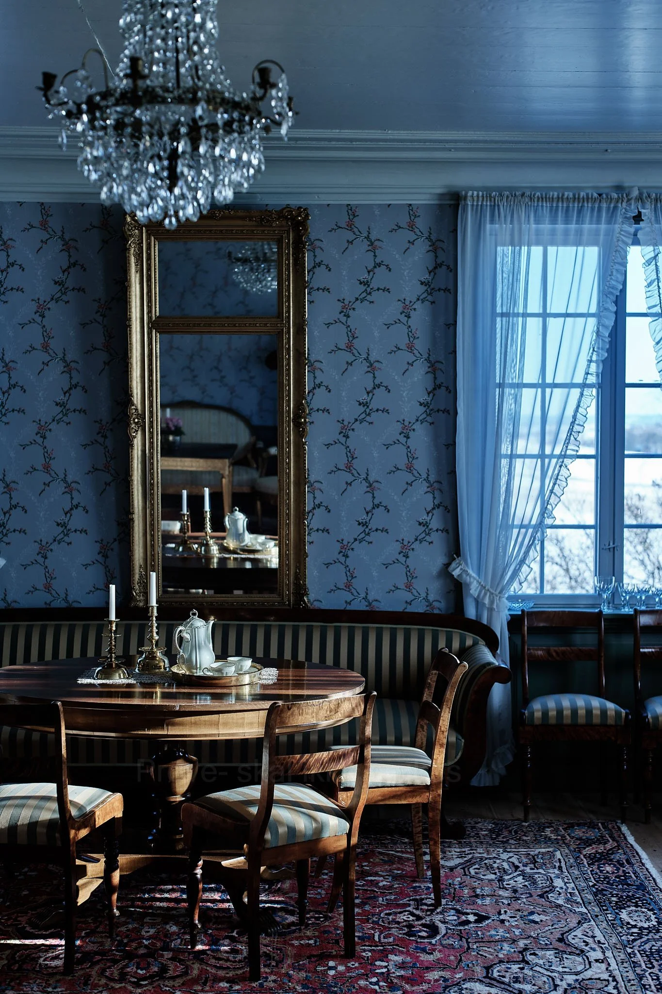 A vintage dining room with a wooden round table, striped upholstered chairs, and a matching curved bench. The table has two tall candlesticks, a ceramic pitcher, and teacups. The room features patterned wallpaper, a large ornate mirror, a window with
