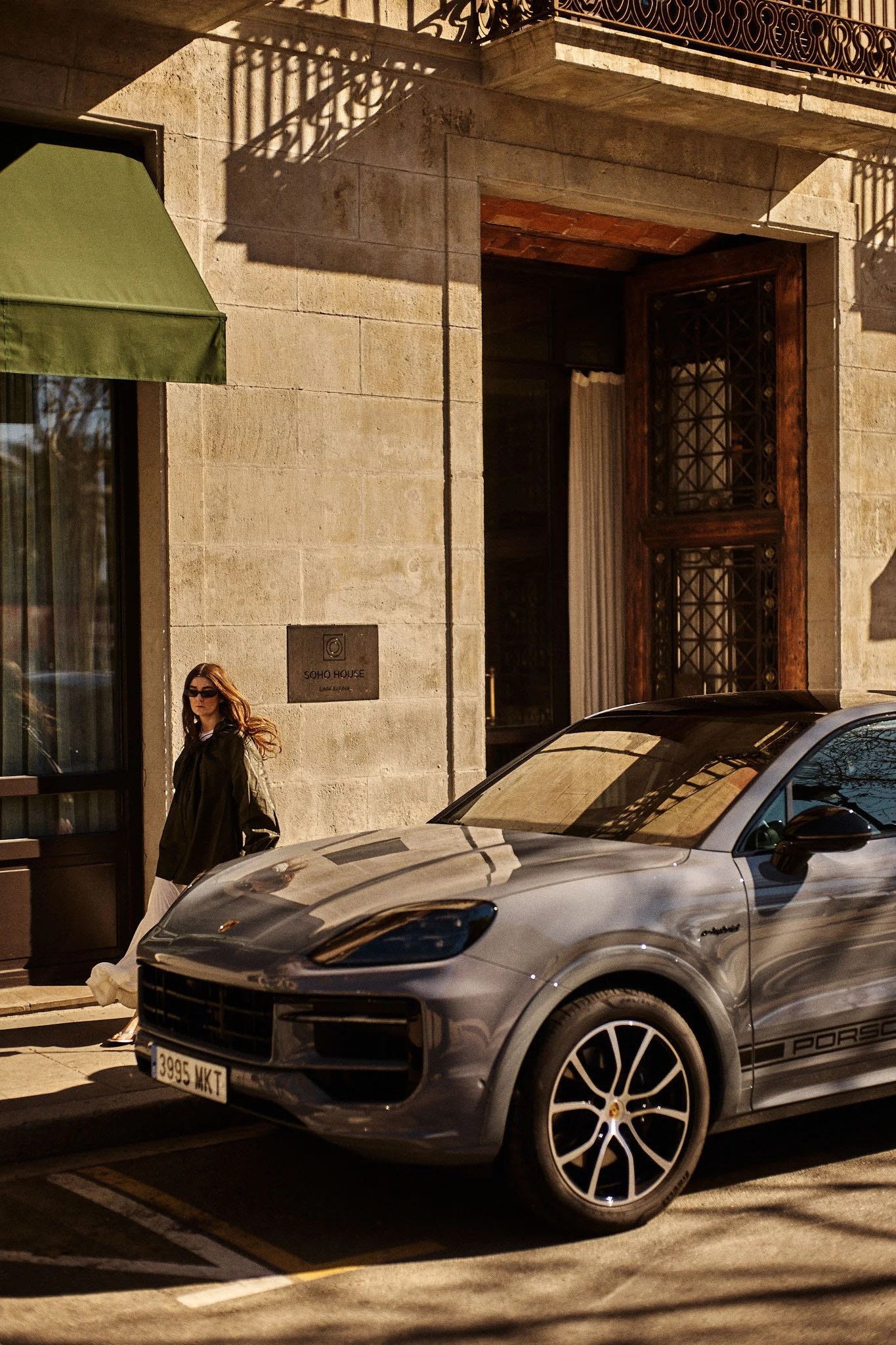 A woman with long hair wearing sunglasses, a dark jacket, and light-colored pants walking beside a parked gray Porsche SUV on a city street, with a stone building and a green awning in the background.