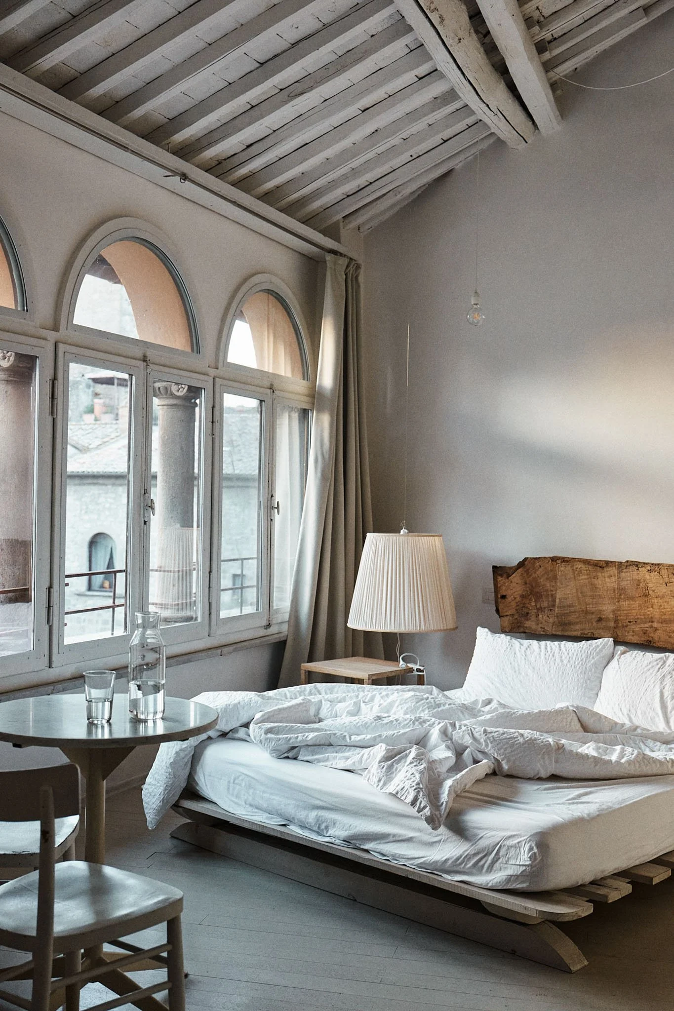 A rustic bedroom with a large window, unmade bed with white linens, a lampshade, wooden headboard, and a small side table with a glass pitcher and two glasses.