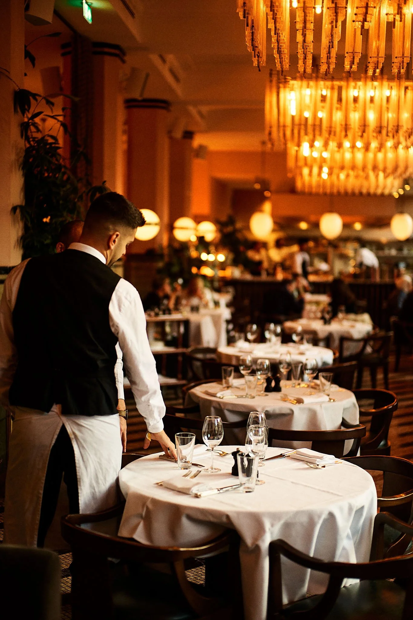 A restaurant waiter adjusting a table set with glasses, silverware, and napkins in a dimly lit, elegant dining room with large chandeliers.