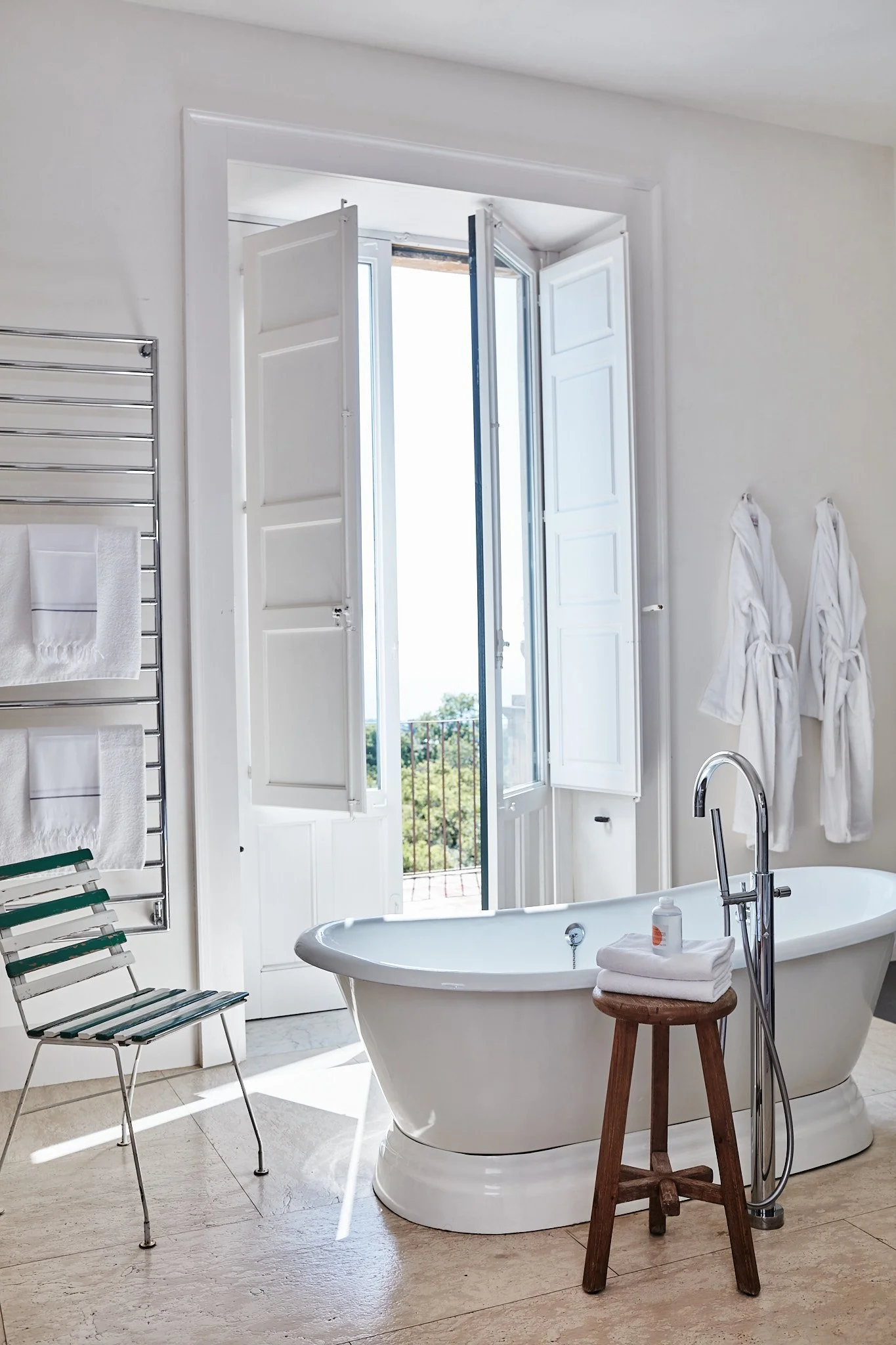 A bright bathroom with a white clawfoot bathtub near an open balcony door, two white robes hanging on the wall, and a small wooden stool with towels and bath products.
