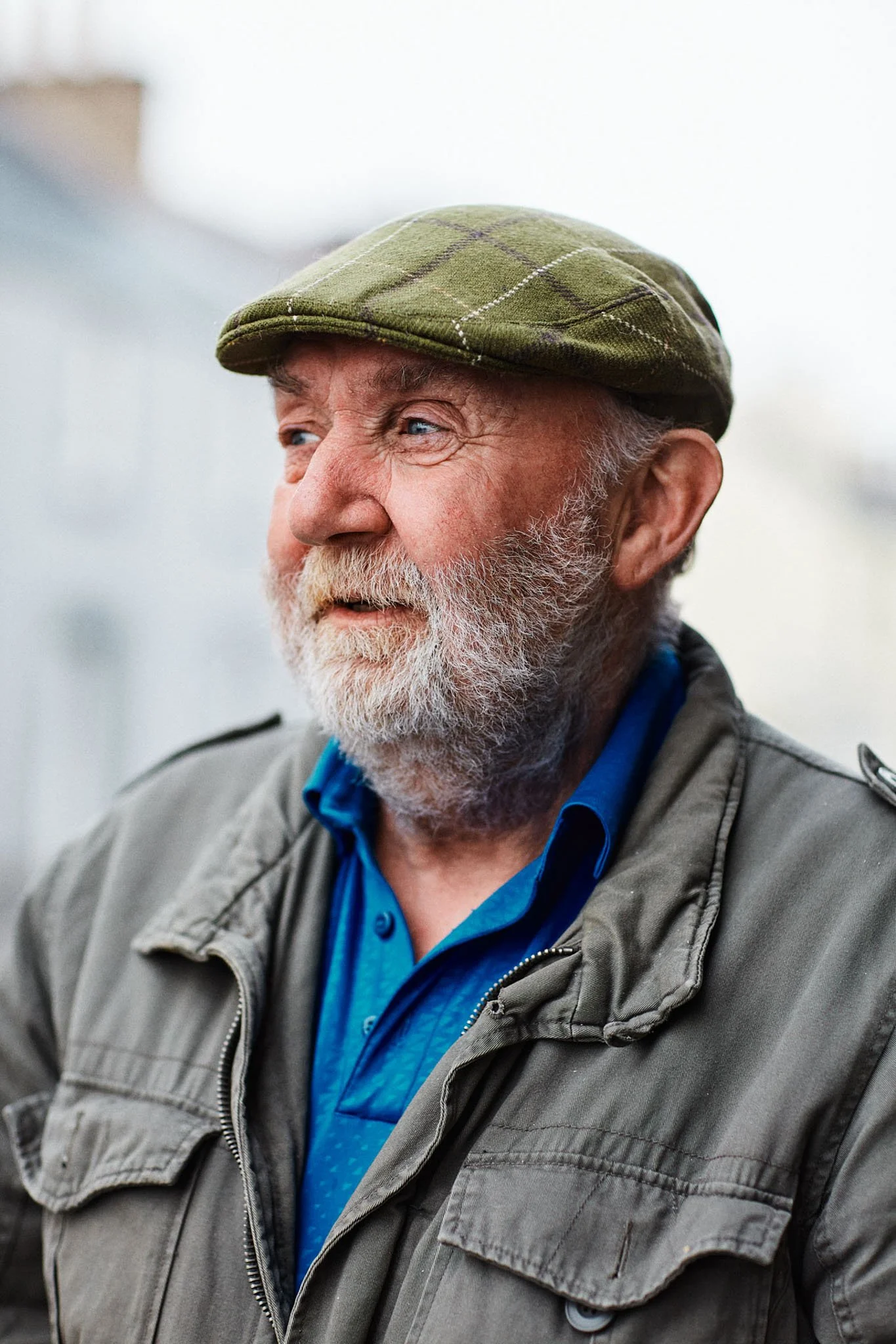 Close-up of an elderly man with a white beard, wearing a green plaid flat cap, a gray jacket, and a blue shirt, outdoors.