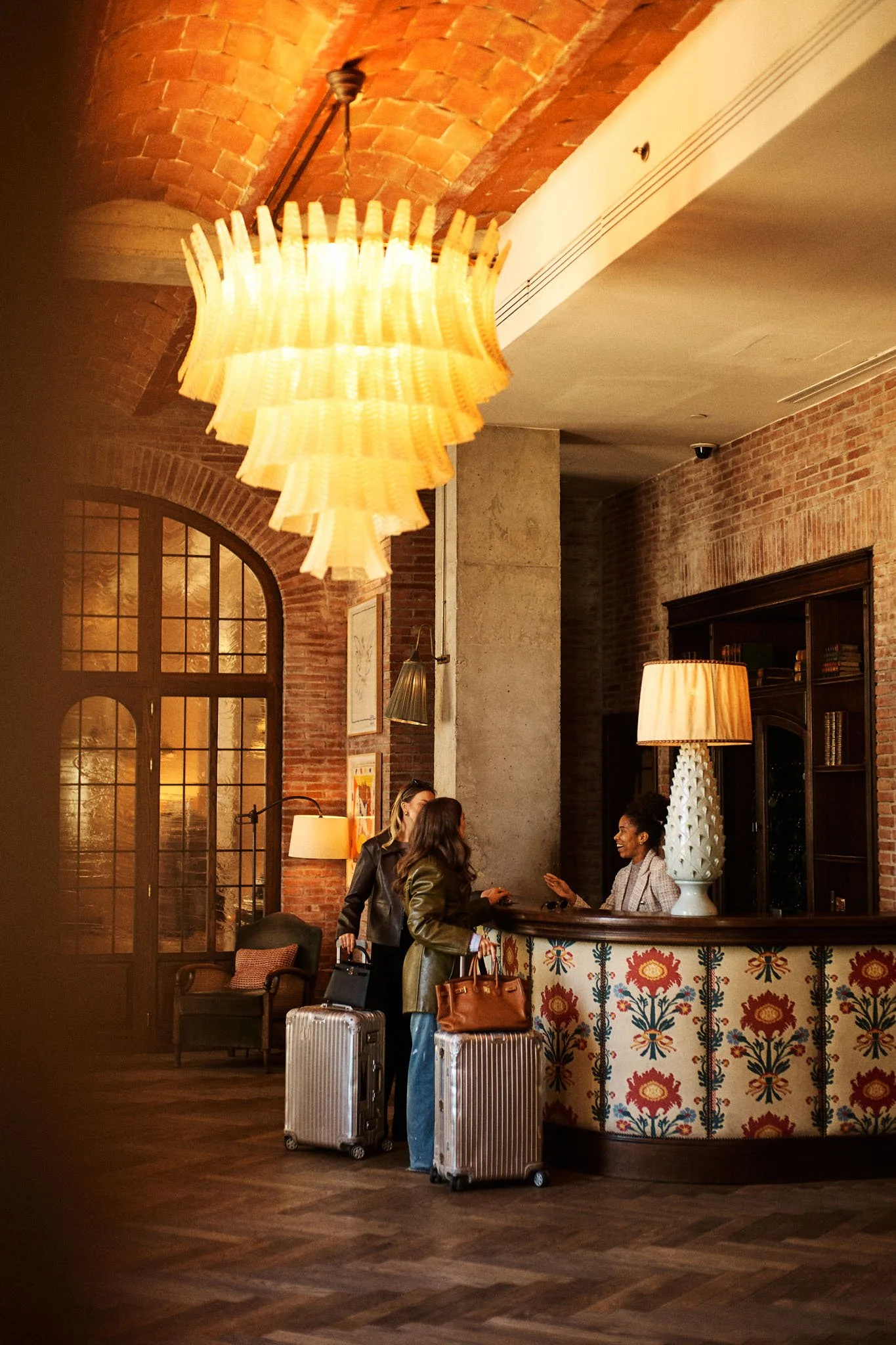 Hotel check-in front desk with two women paying, carrying luggage, in a warm, brick, vintage-style lobby.