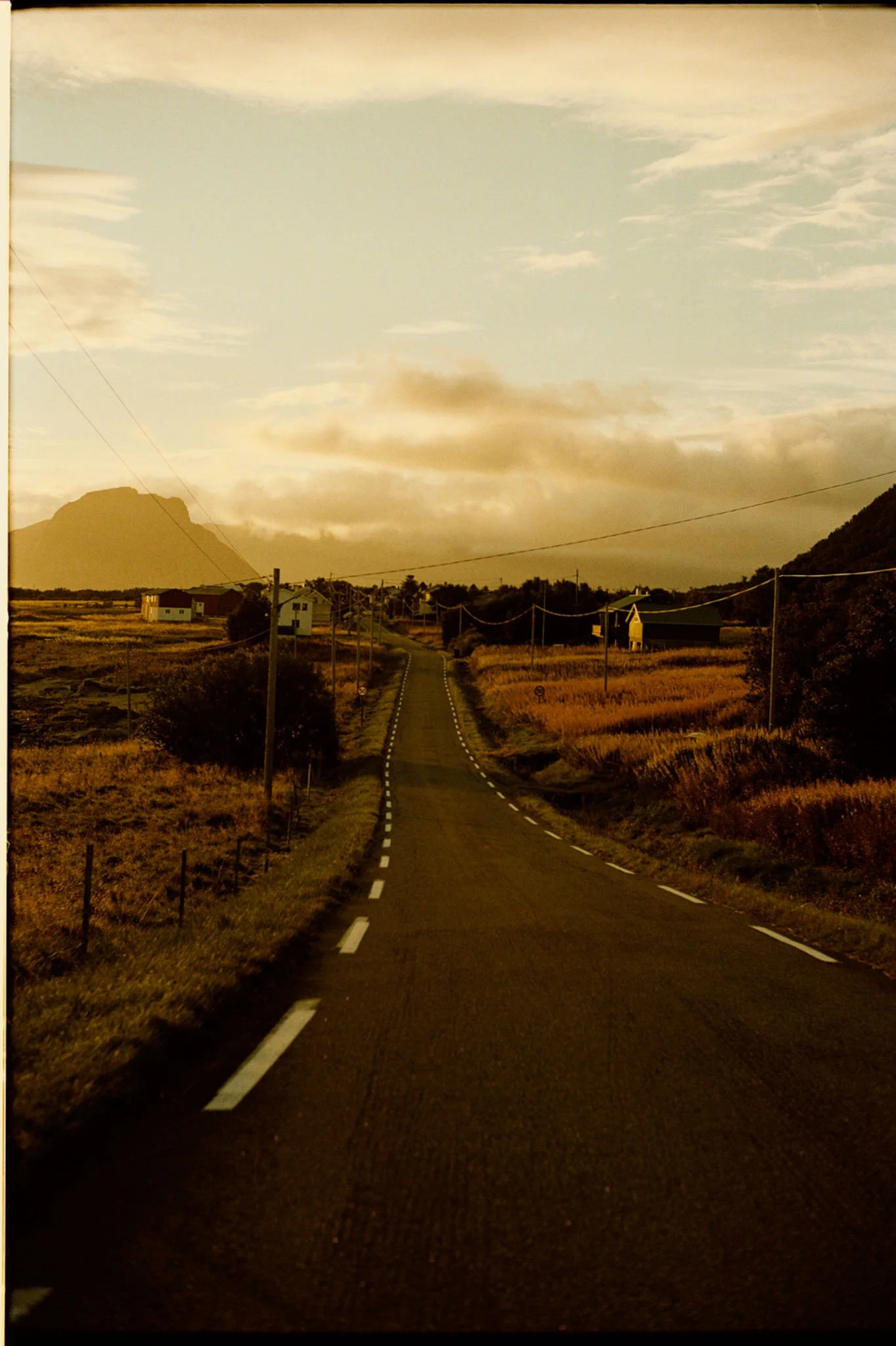 A long, winding rural road with dashed white lines, passing through open fields with some houses and utility poles on either side, leading toward mountains in the background under a sky with clouds during sunset or sunrise.