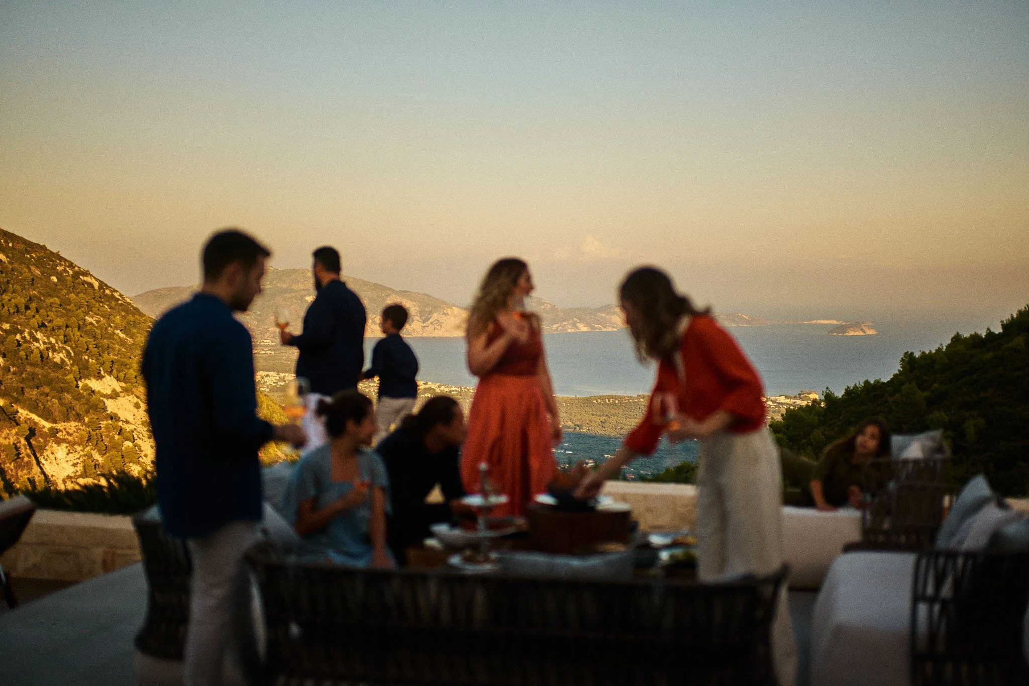 People enjoying a gathering outdoors with a scenic view of hills, ocean, and sky at sunset.