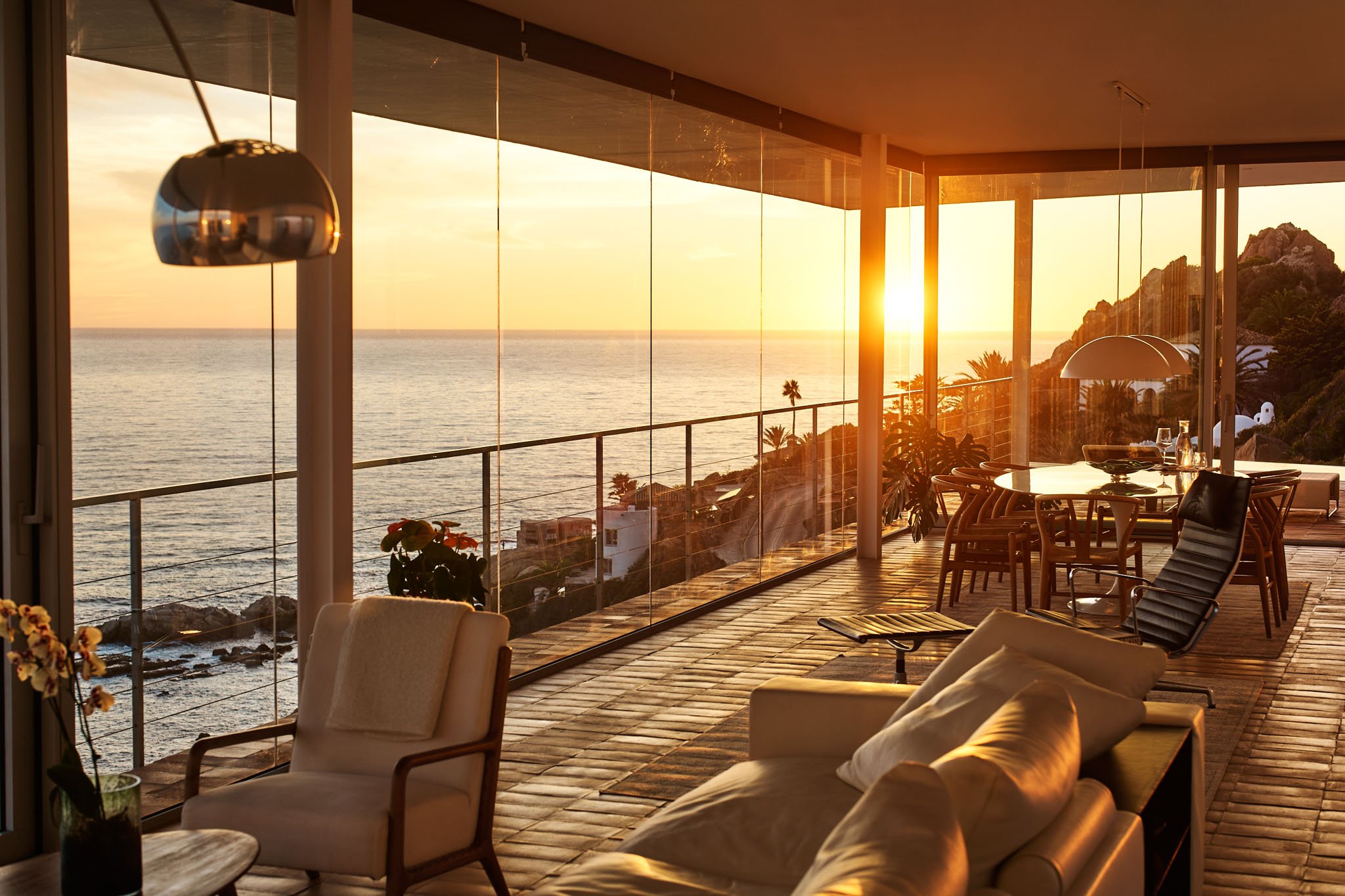 Living room with large glass windows showing an ocean view at sunset, with a dining table, chairs, lounge chairs, and decorative plants.