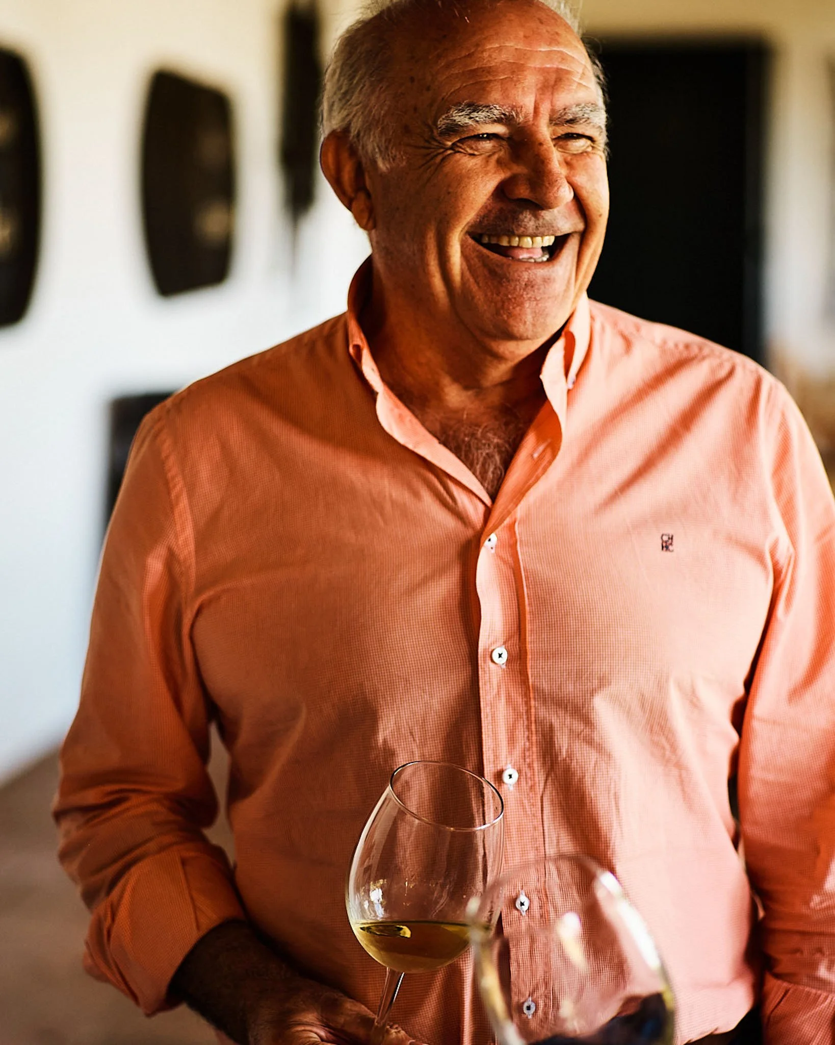 An elderly man with gray hair and a broad smile, wearing a peach-colored button-up shirt, holding a glass of white wine, standing indoors in warm lighting.