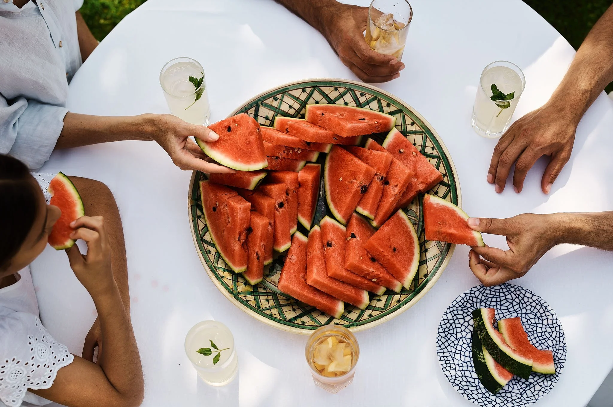 People gathered around a table with a large bowl of sliced watermelon, some in a blue patterned bowl, and glasses of drinks including lemonade and iced tea, outdoors.