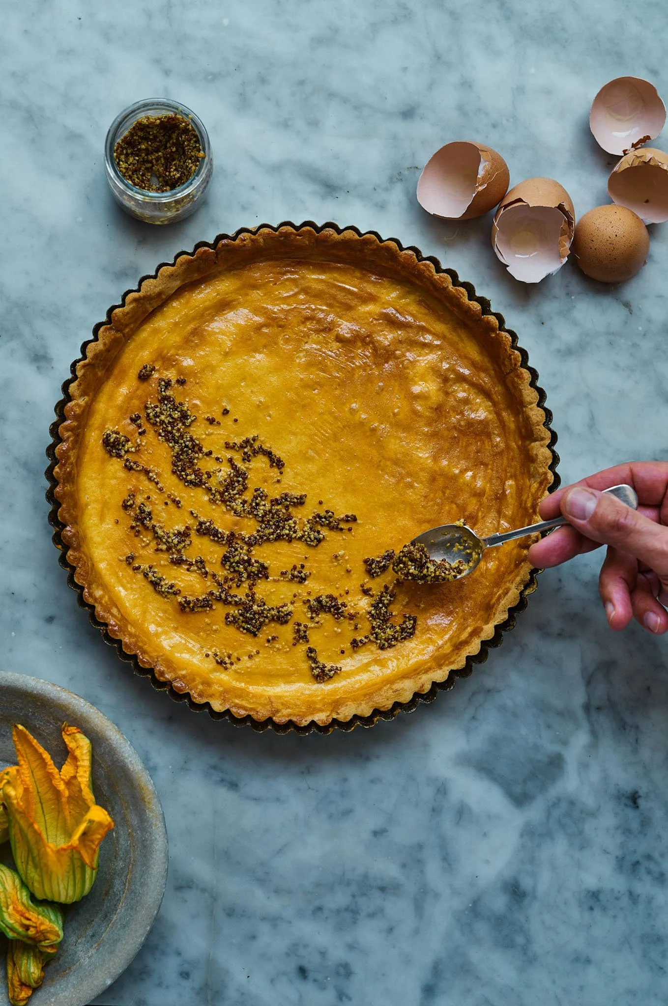 A partially prepared lemon tart with black mustard seeds on top, on a marble countertop, with eggshells and quail eggs nearby.