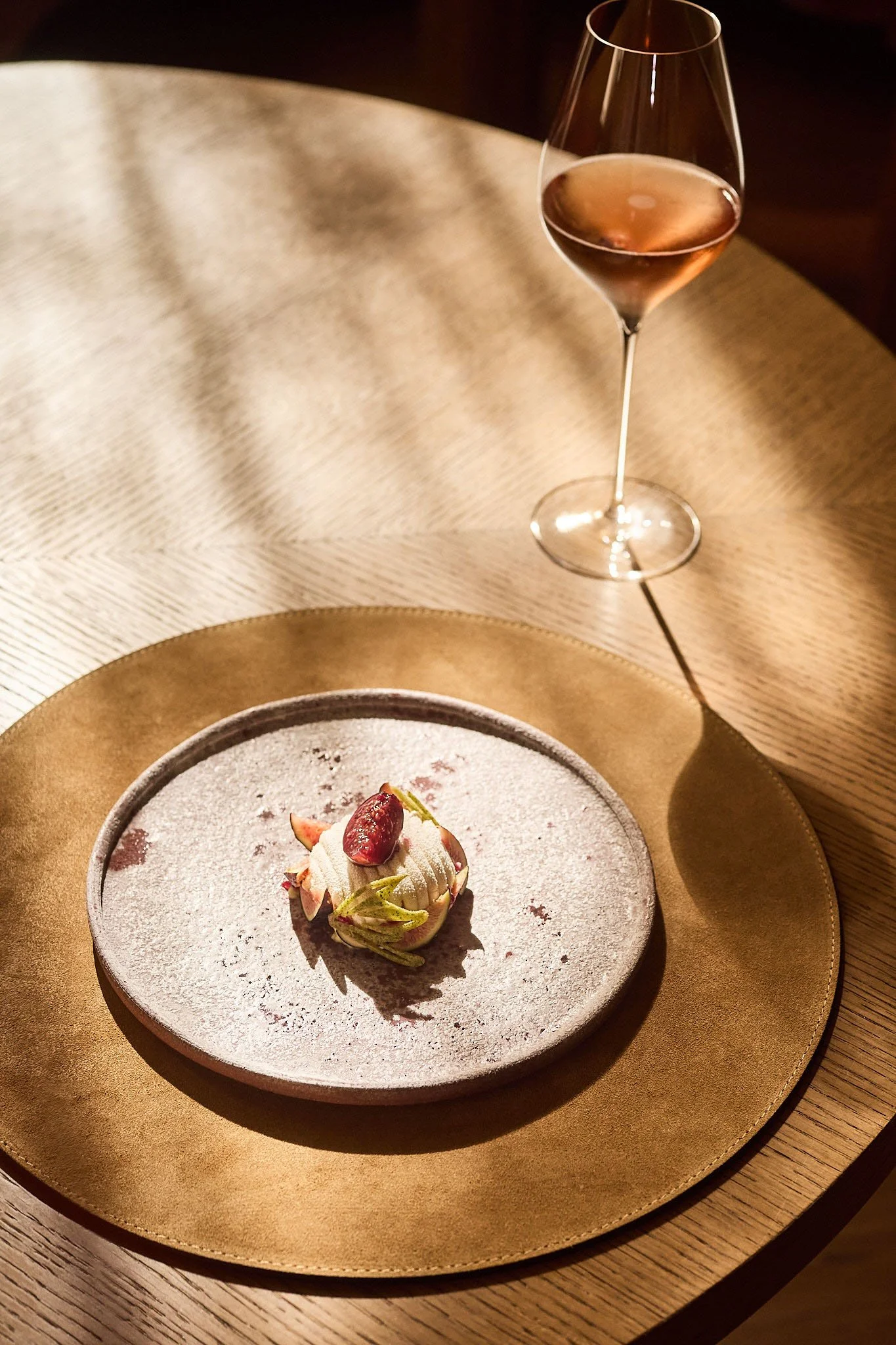 A dessert topped with a strawberry and mint on a round plate, a glass of pink wine on a table with a gold placemat and textured beige cloth.