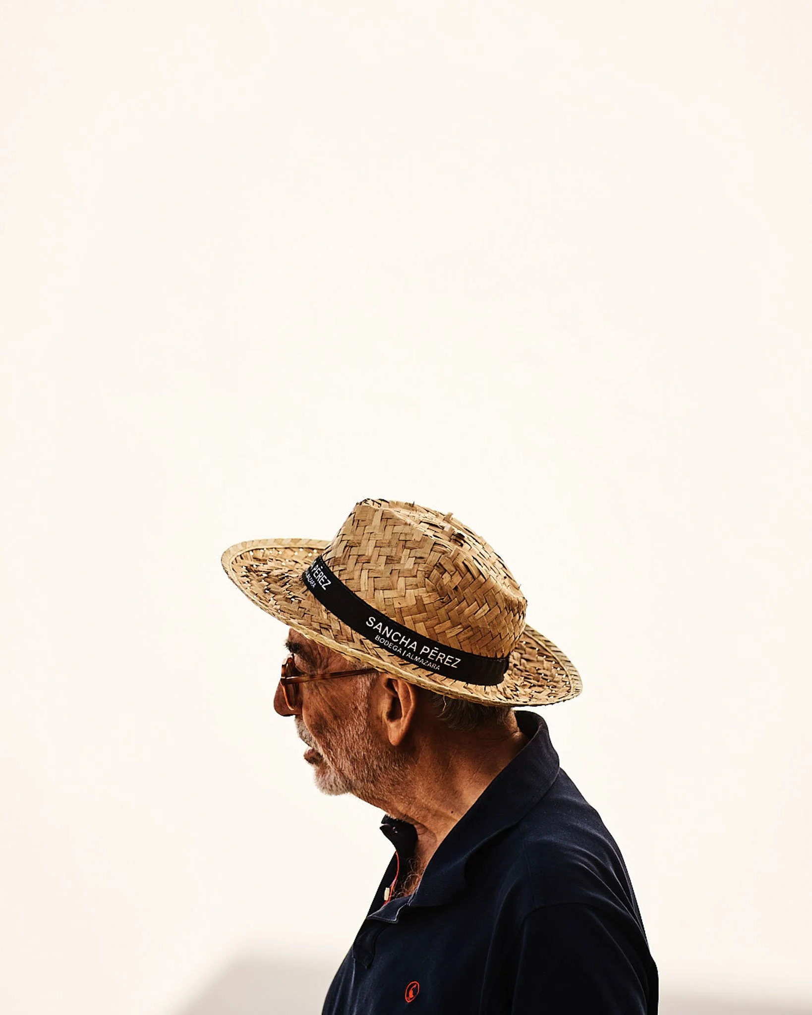 Side view of an older man wearing sunglasses, a navy polo shirt, and a large straw hat with a black band, standing against a plain off-white background.
