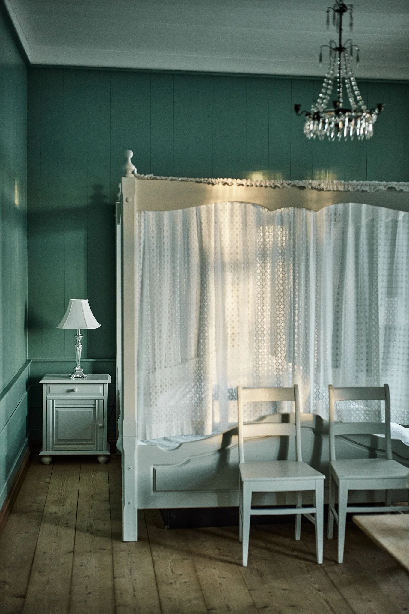 A bedroom with green walls, a white four-poster bed with curtains, two white chairs in front, a white nightstand with a lamp, a chandelier hanging from the ceiling, and wooden floor.
