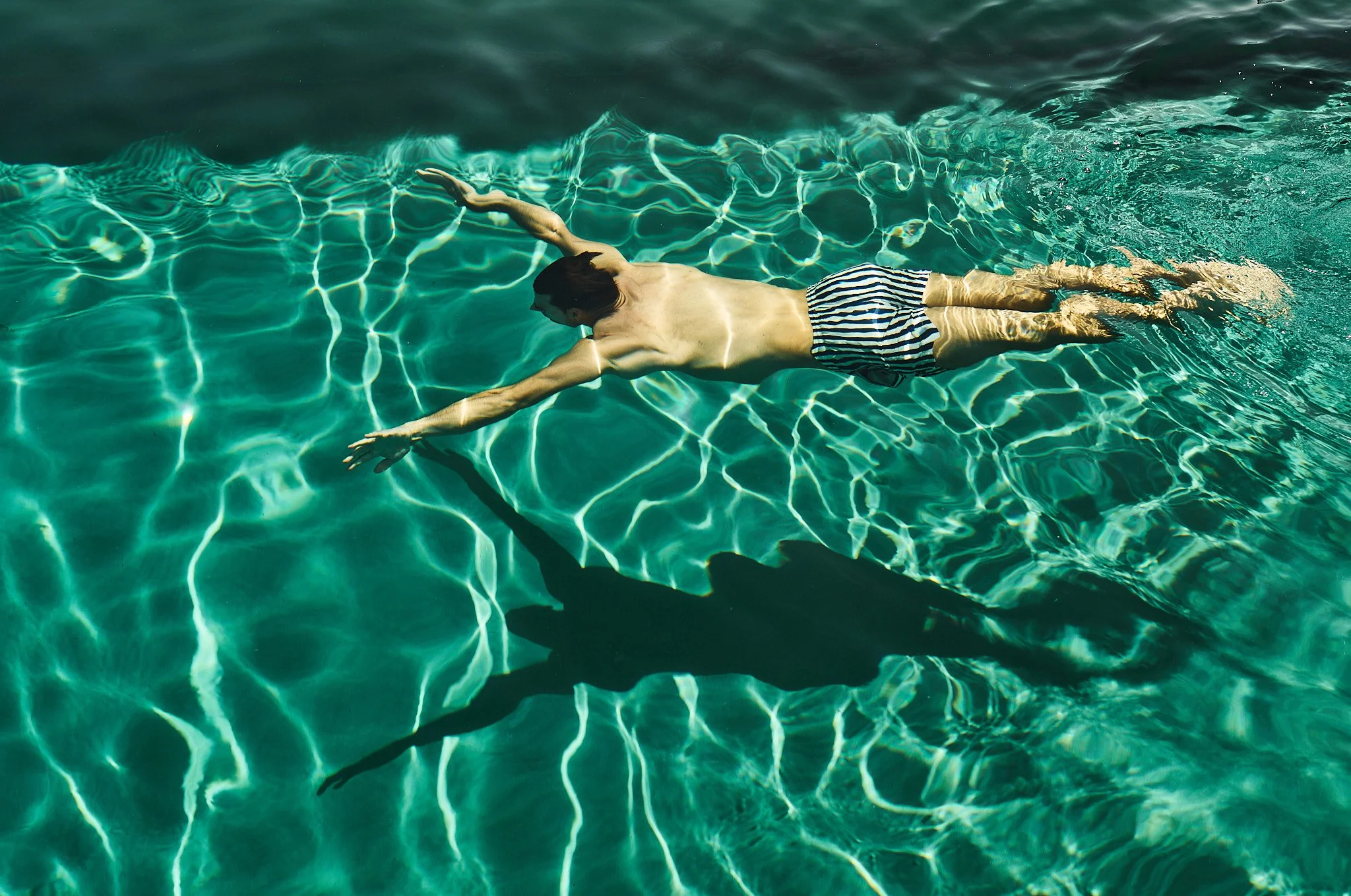 A man swimming in clear water, wearing striped swim trunks, with his arms extended forward and casting a shadow on the water's surface.