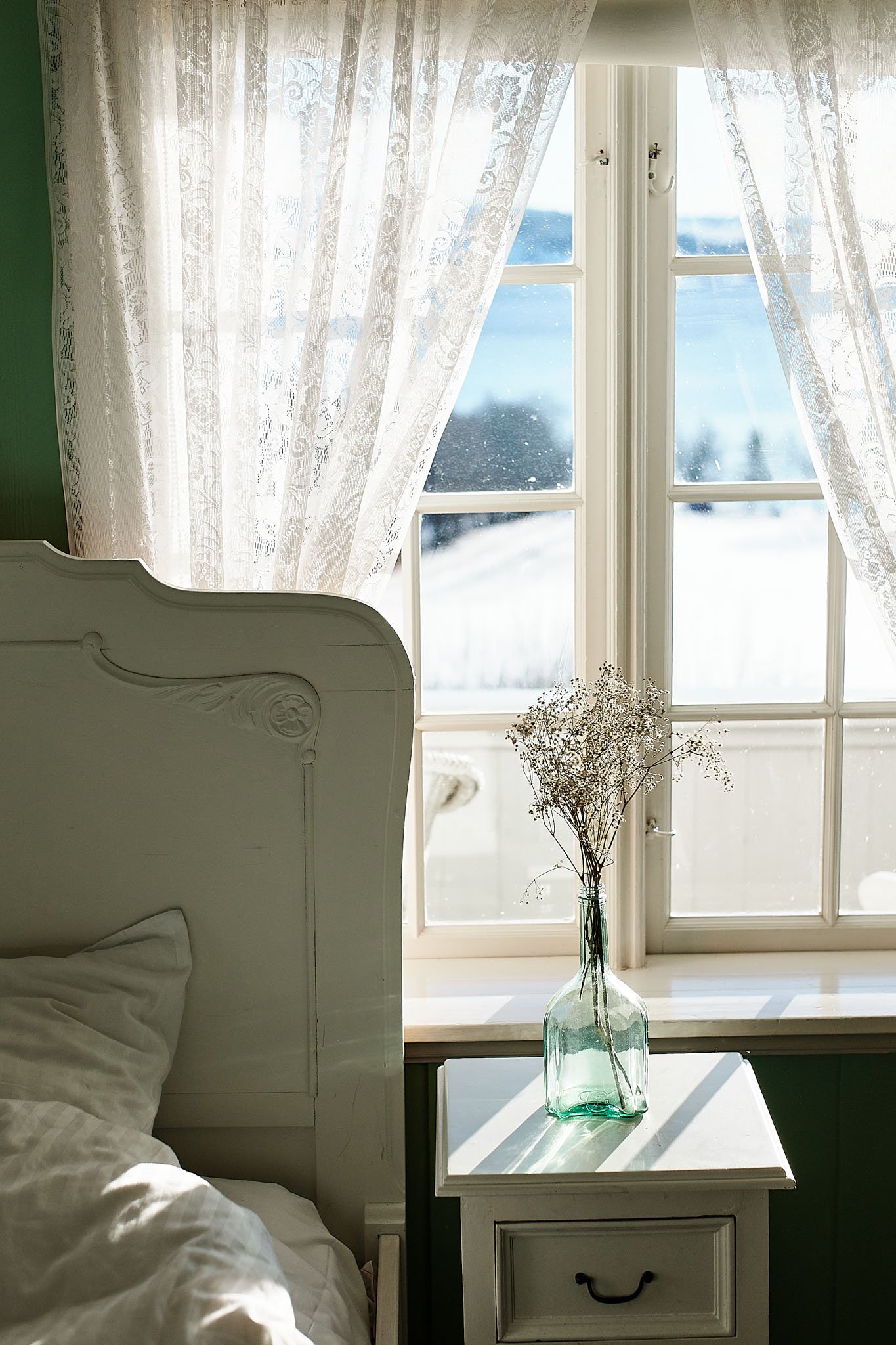 A cozy bedroom with a white bed, a nightstand with a glass vase and dried flowers, and a large window with lace curtains showing a snowy outdoor landscape.