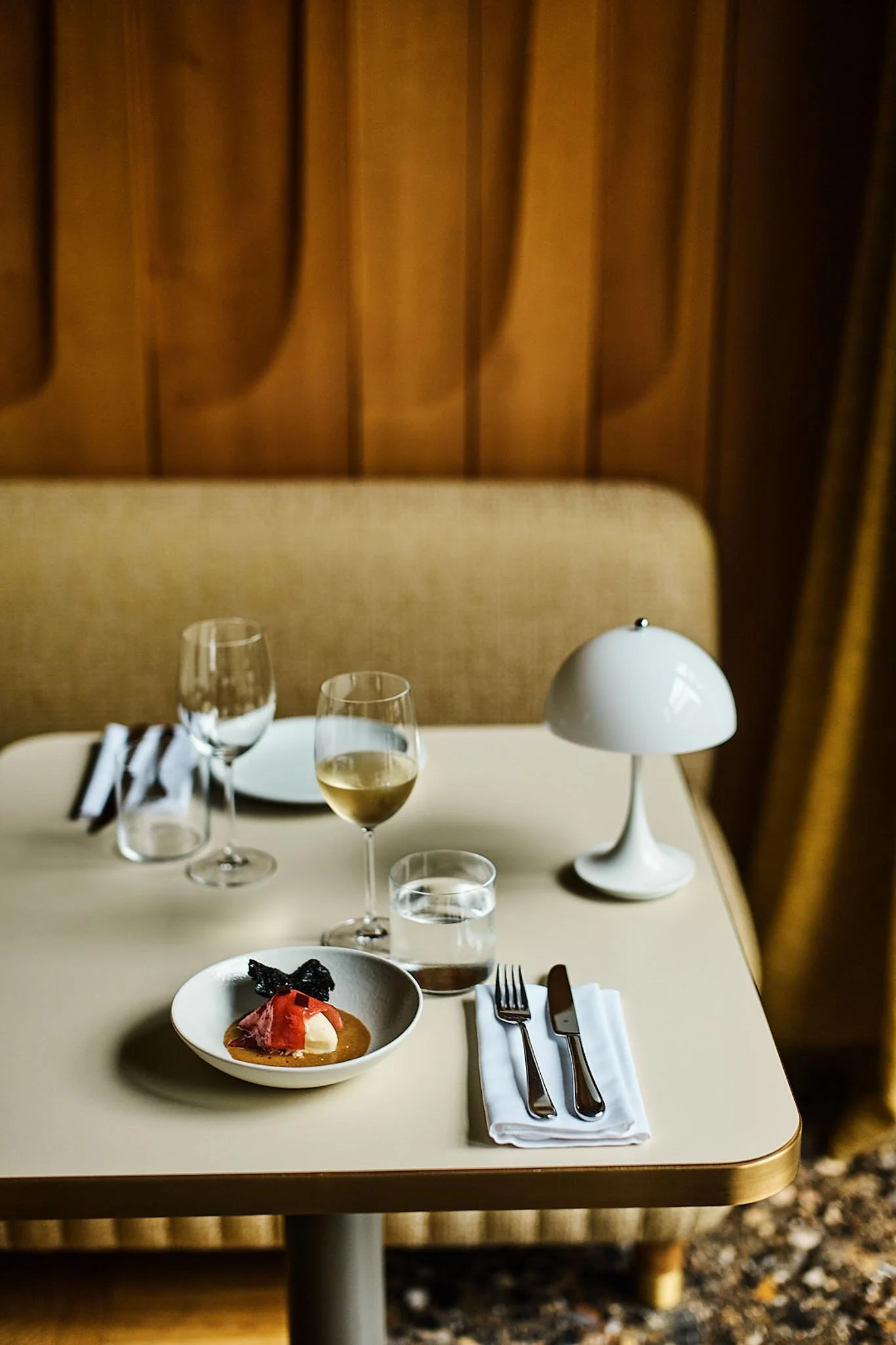 A dining table set with a white plate, a dessert, a glass of white wine, a glass of water, an empty wine glass, a table lamp, and silverware on a white napkin, with a beige cushioned booth and wooden wall in the background.