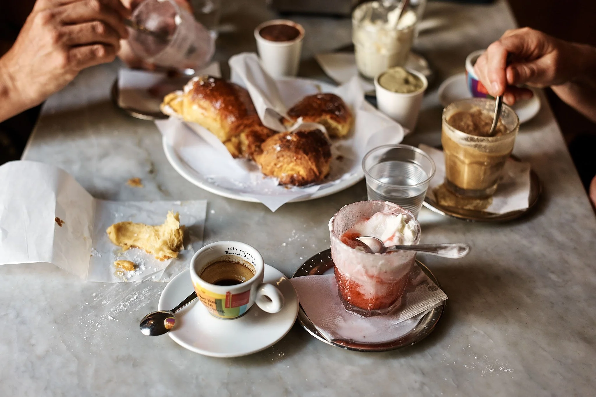 Table with various breakfast items, including a croissant, jam, yogurt, coffee, and other condiments, with two people reaching for food.
