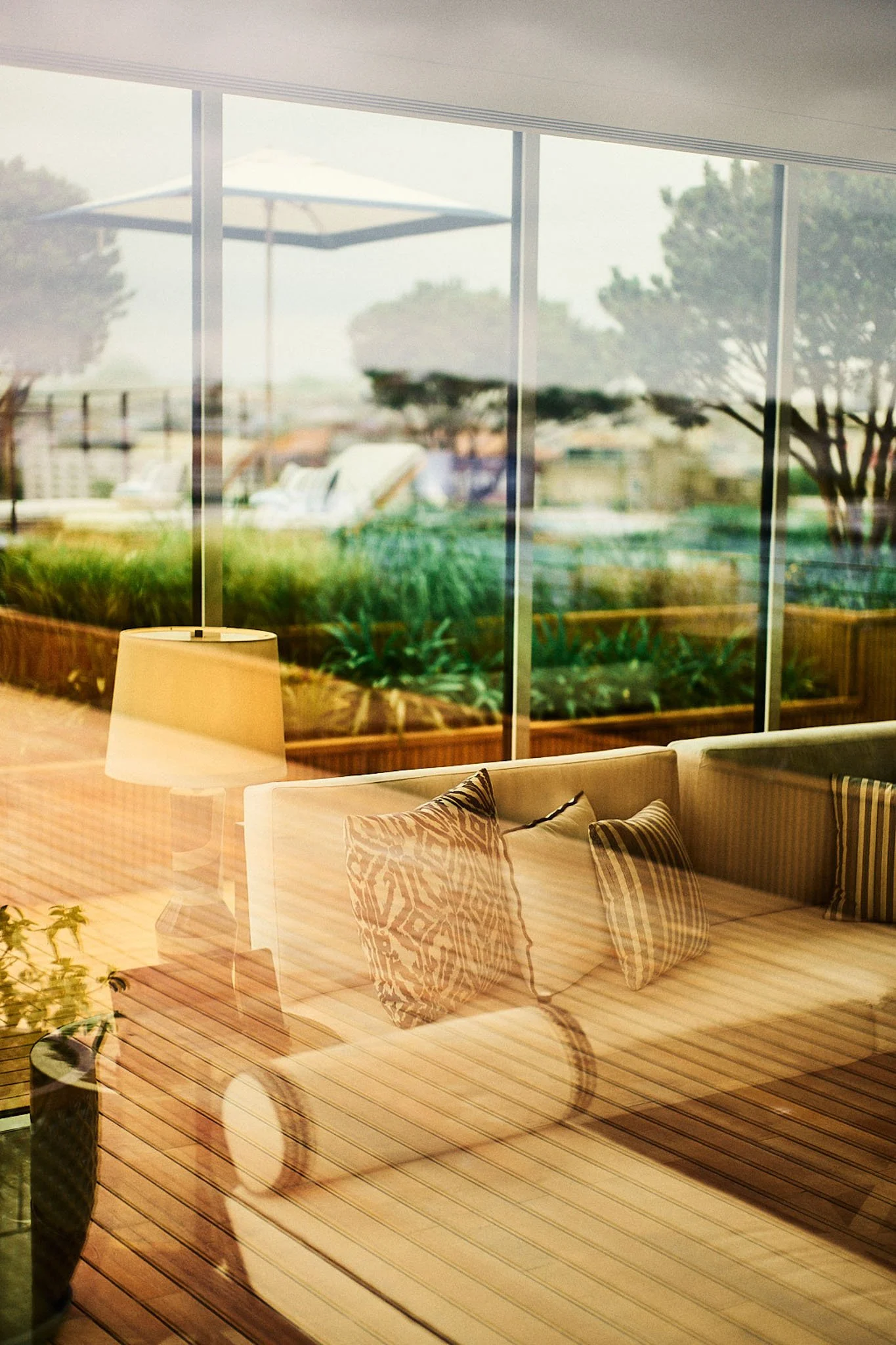 Interior of a living room with a beige couch, decorative pillows, a wooden coffee table, a table lamp, and a large window showing outdoor patio furniture and greenery.