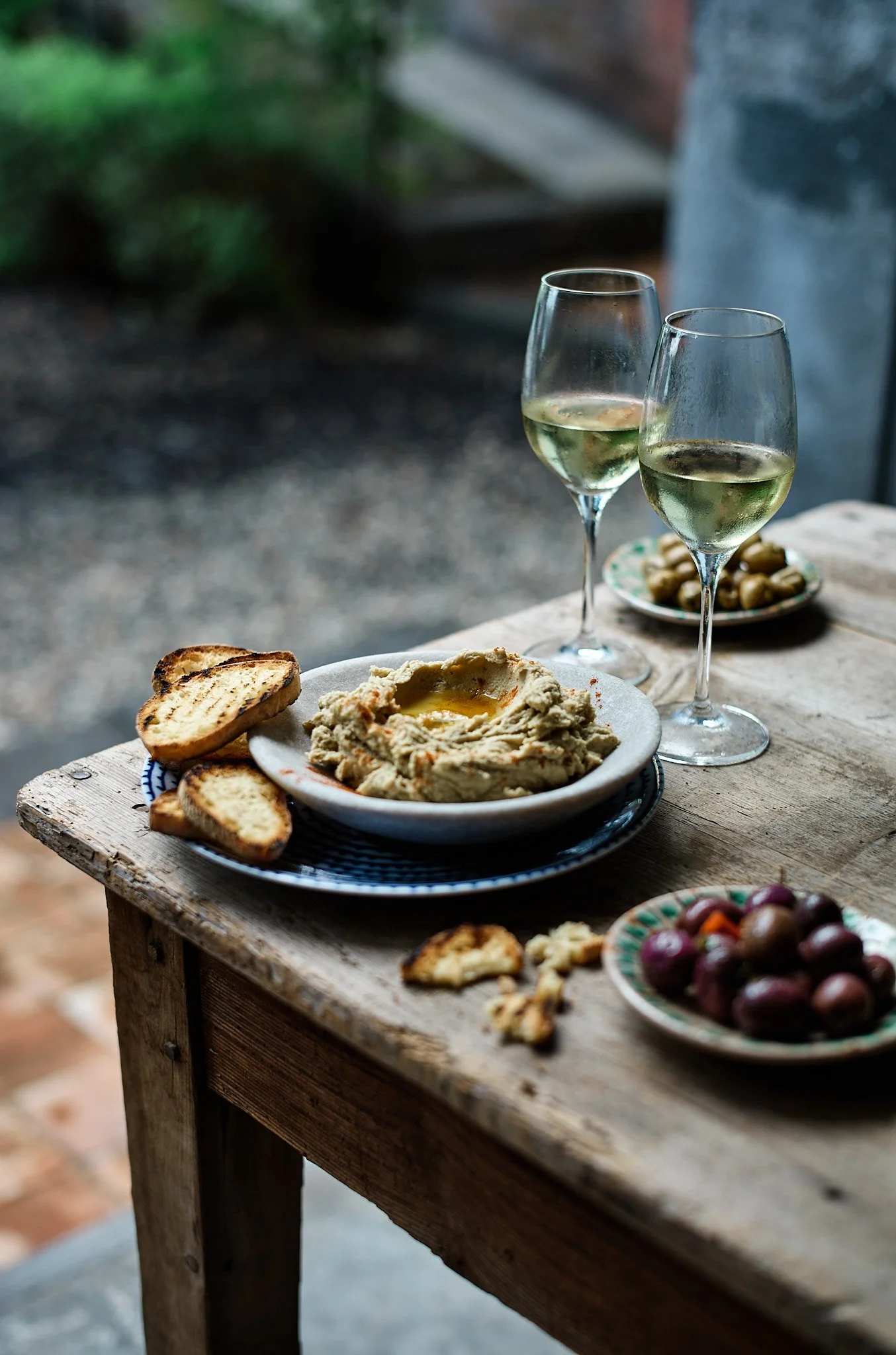 A rustic wooden table with a plate of hummus, toasted bread slices, two glasses of white wine, and small bowls of grapes and olives.
