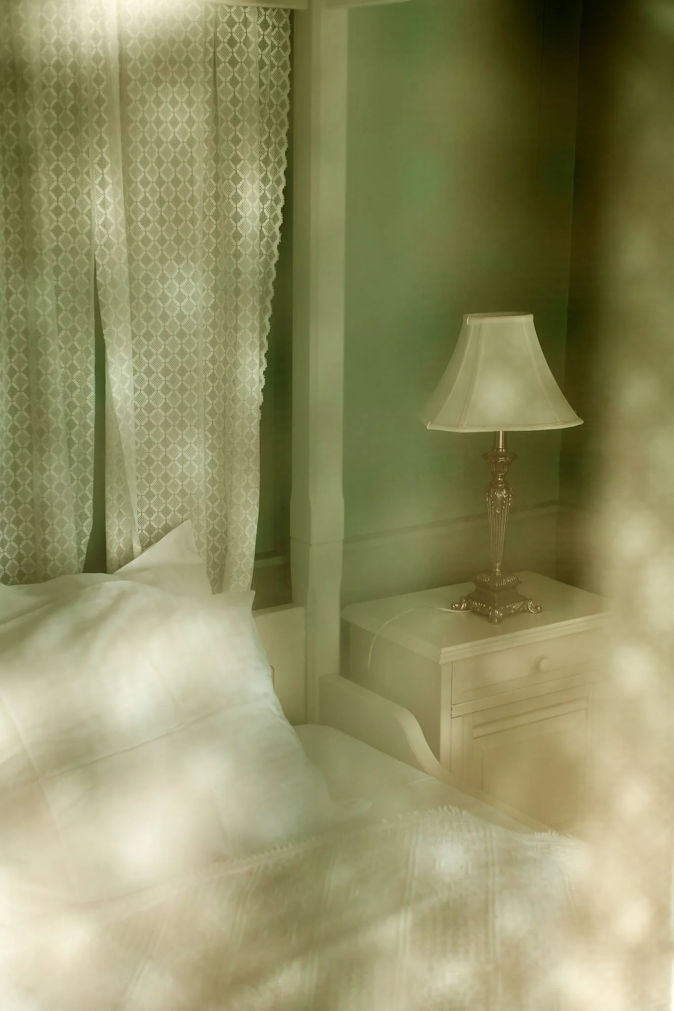 A bedroom with a white bed, a nightstand with a lamp, and curtains on a green wall.