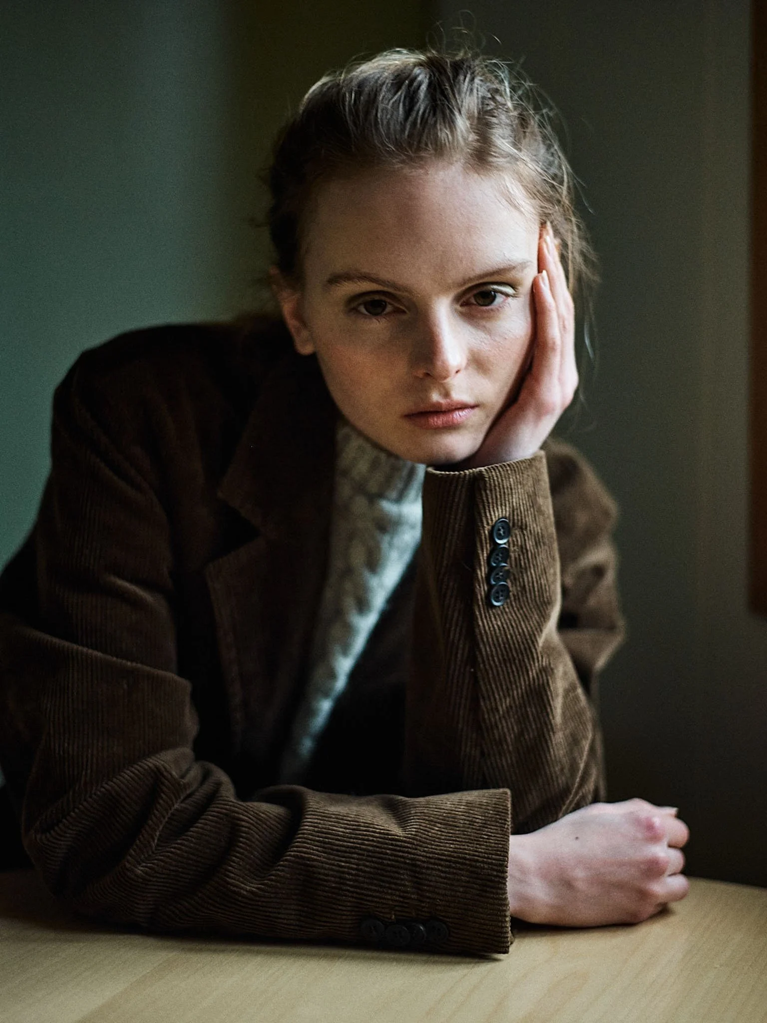 A young woman with blonde hair, wearing a brown corduroy jacket and a cream-colored sweater, resting her head on her hand and looking at the camera while seated at a table.