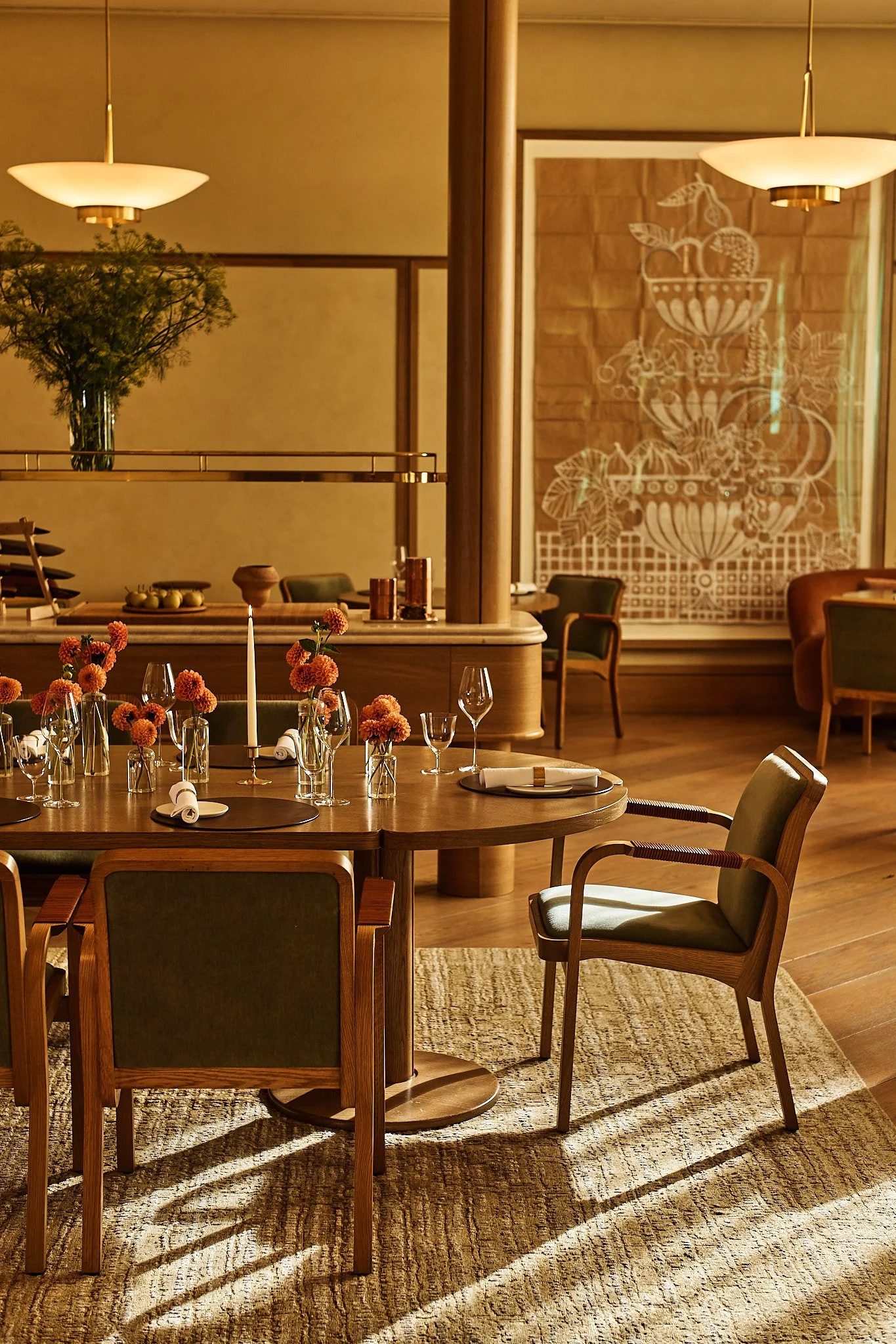 A elegantly decorated restaurant interior with a wooden table set with orange flowers in glass vases, wine glasses, and napkins. The room features warm lighting, large lamps, potted plants, and wall art with a fruit pattern, creating a cozy, sophisti