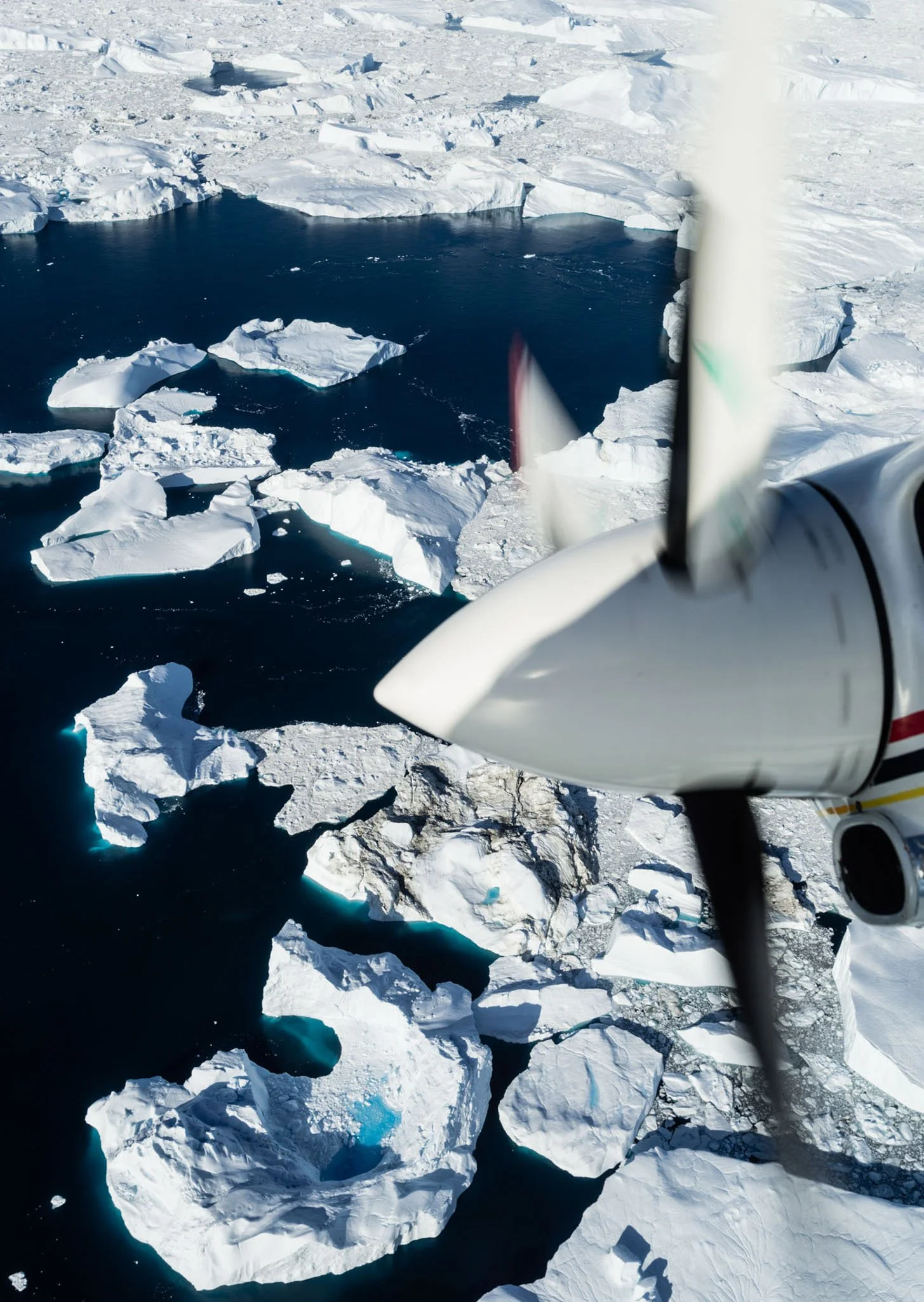 View from a small aircraft showing floating icebergs and ice-covered water in a polar region.