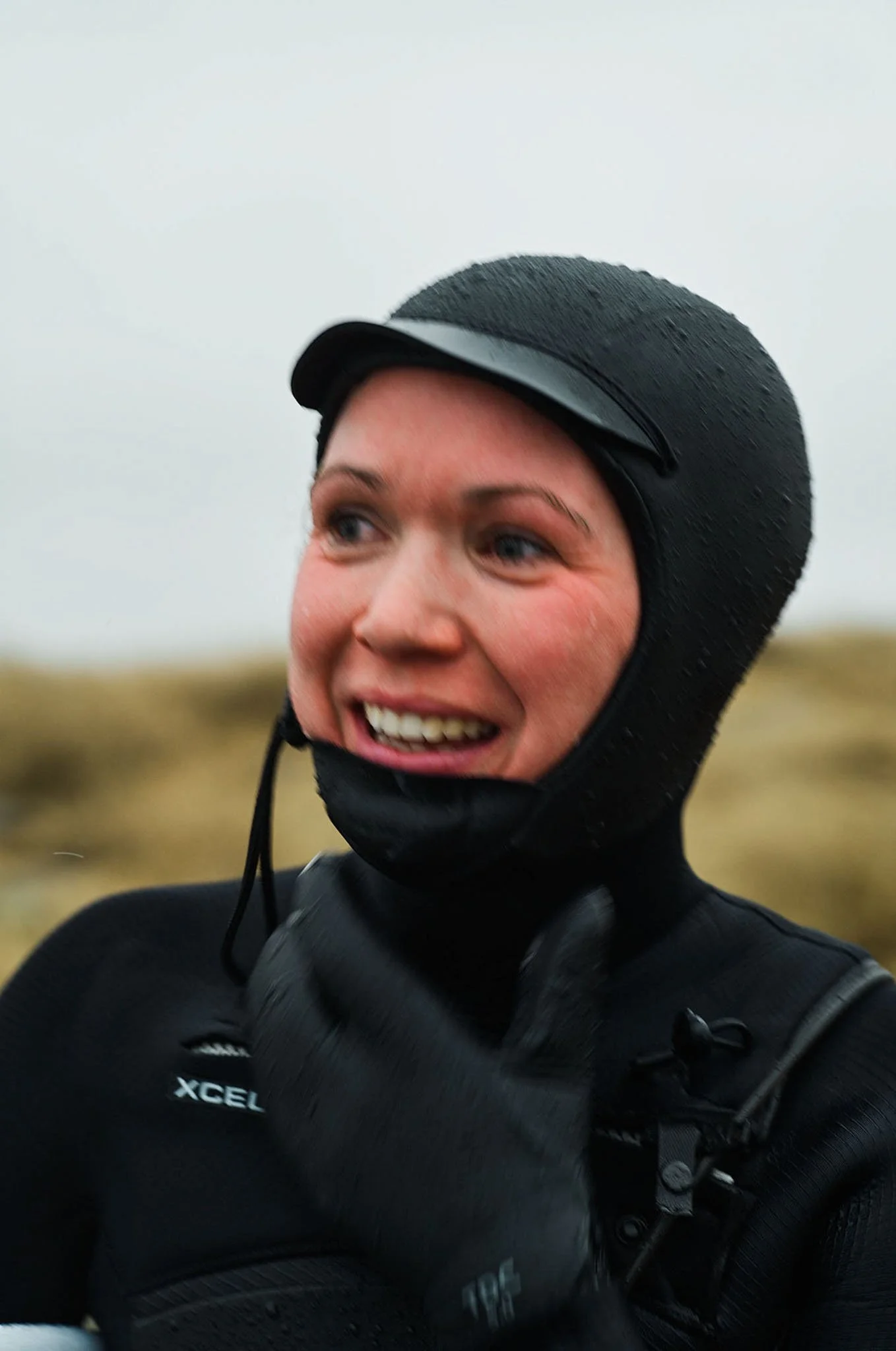 A woman wearing a black helmet and black gear, smiling outdoors on a cloudy day with a blurred natural landscape in the background.