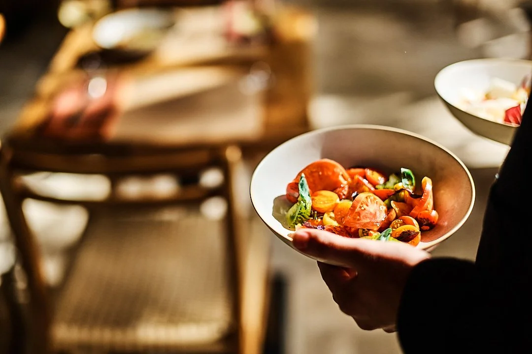 A person holding a white bowl of colorful tomato and basil salad at a dining table.