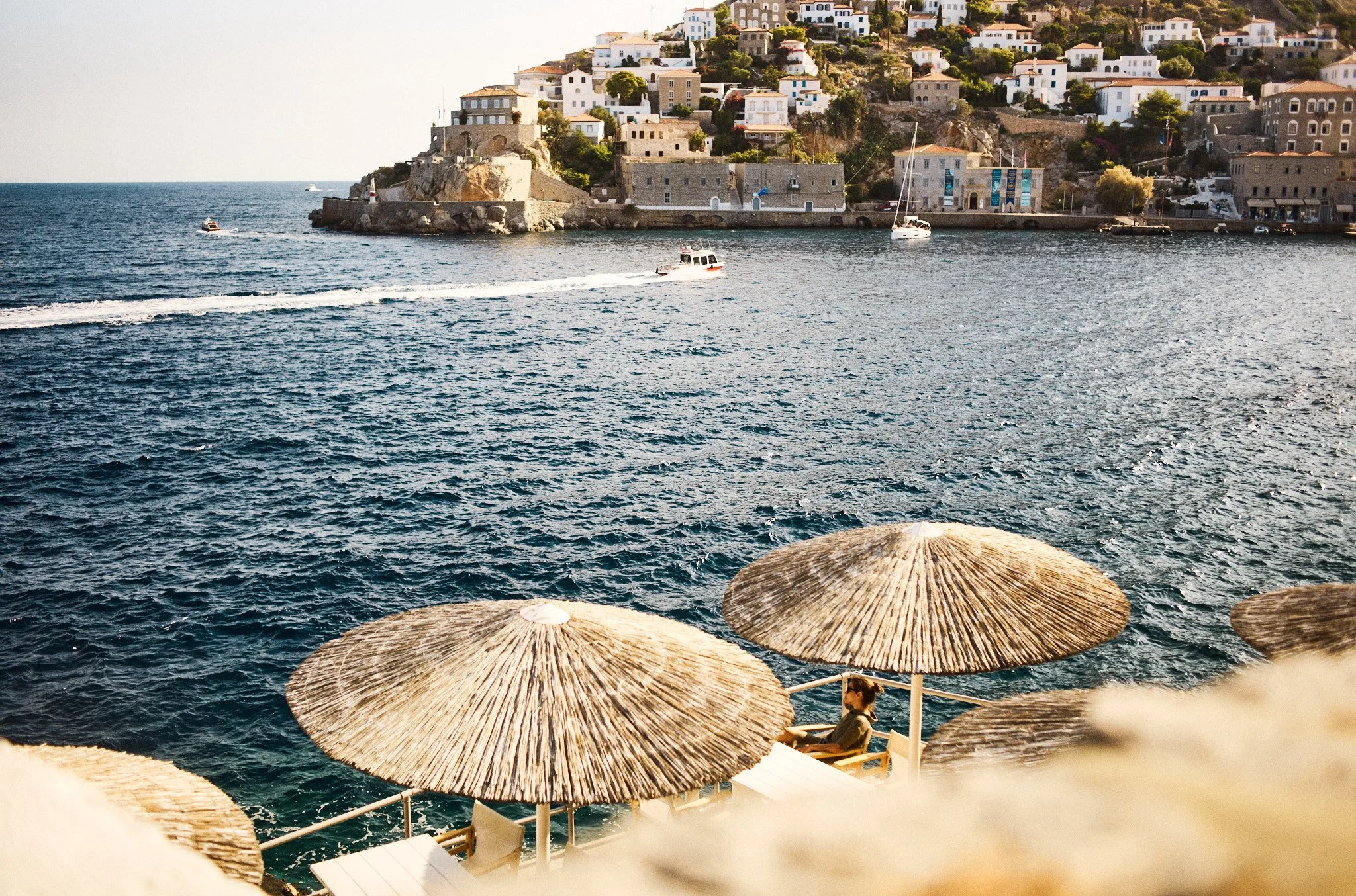 A seaside view with a woman sitting under a straw umbrella on a beach, overlooking the water with boats and hillside houses in the background.
