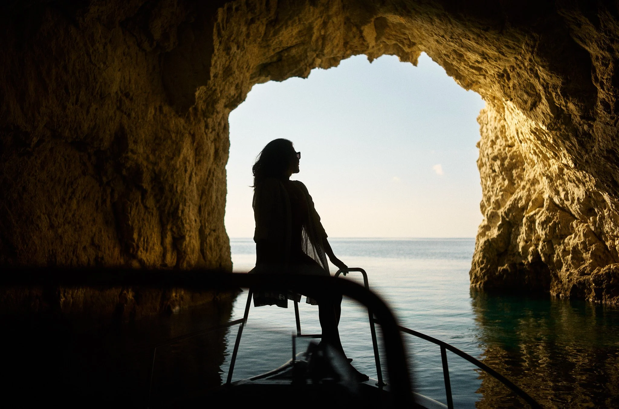 Silhouette of a woman sitting on a boat inside a rocky sea cave, looking out at the ocean and sky.