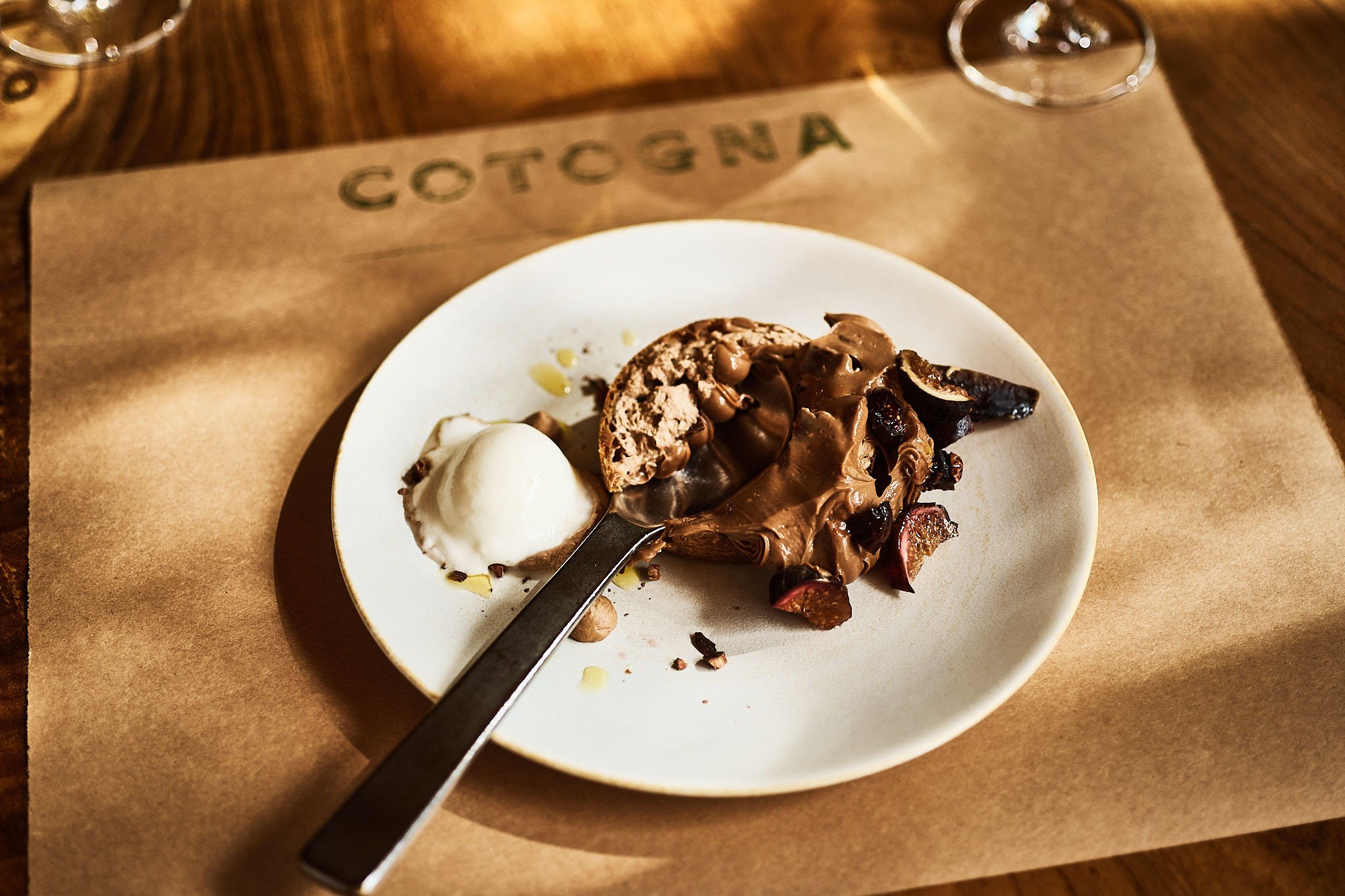 A white plate with chocolate gelato, a chocolate tart or cake, and a scoop of vanilla ice cream, served on a brown paper mat with 'COTOGNA' printed on it, on a wooden table.