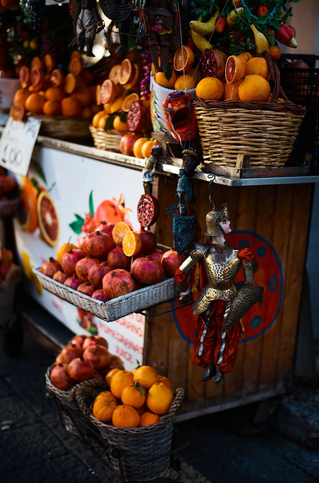 A street market stand with baskets and piles of oranges and pomegranates, decorated with colorful hanging puppets and a painted sign.
