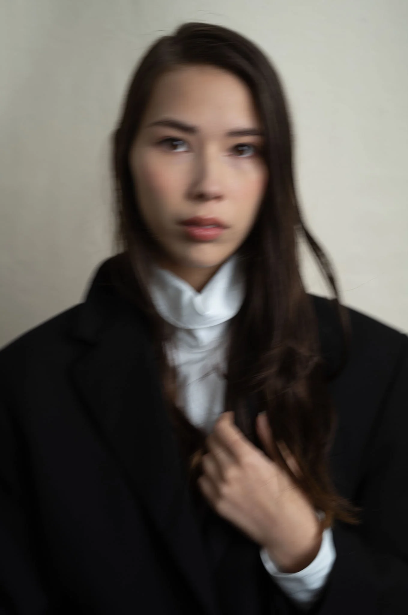 Blurry portrait of a woman with dark hair wearing a black blazer and white shirt, looking at the camera with a neutral expression.