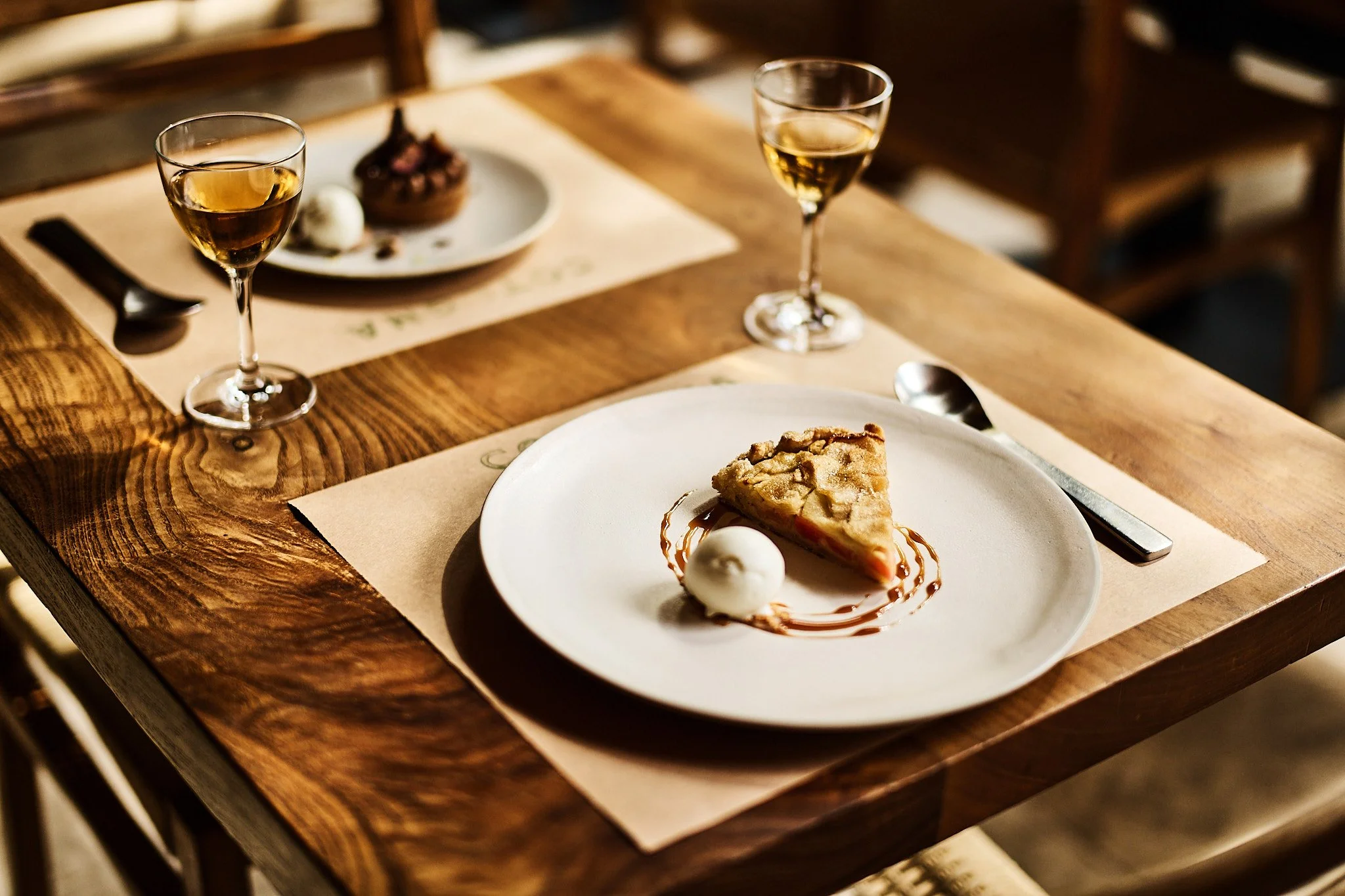 A dessert plate with a slice of pie, a scoop of vanilla ice cream, and caramel sauce, with two glasses of white wine on a wooden table.