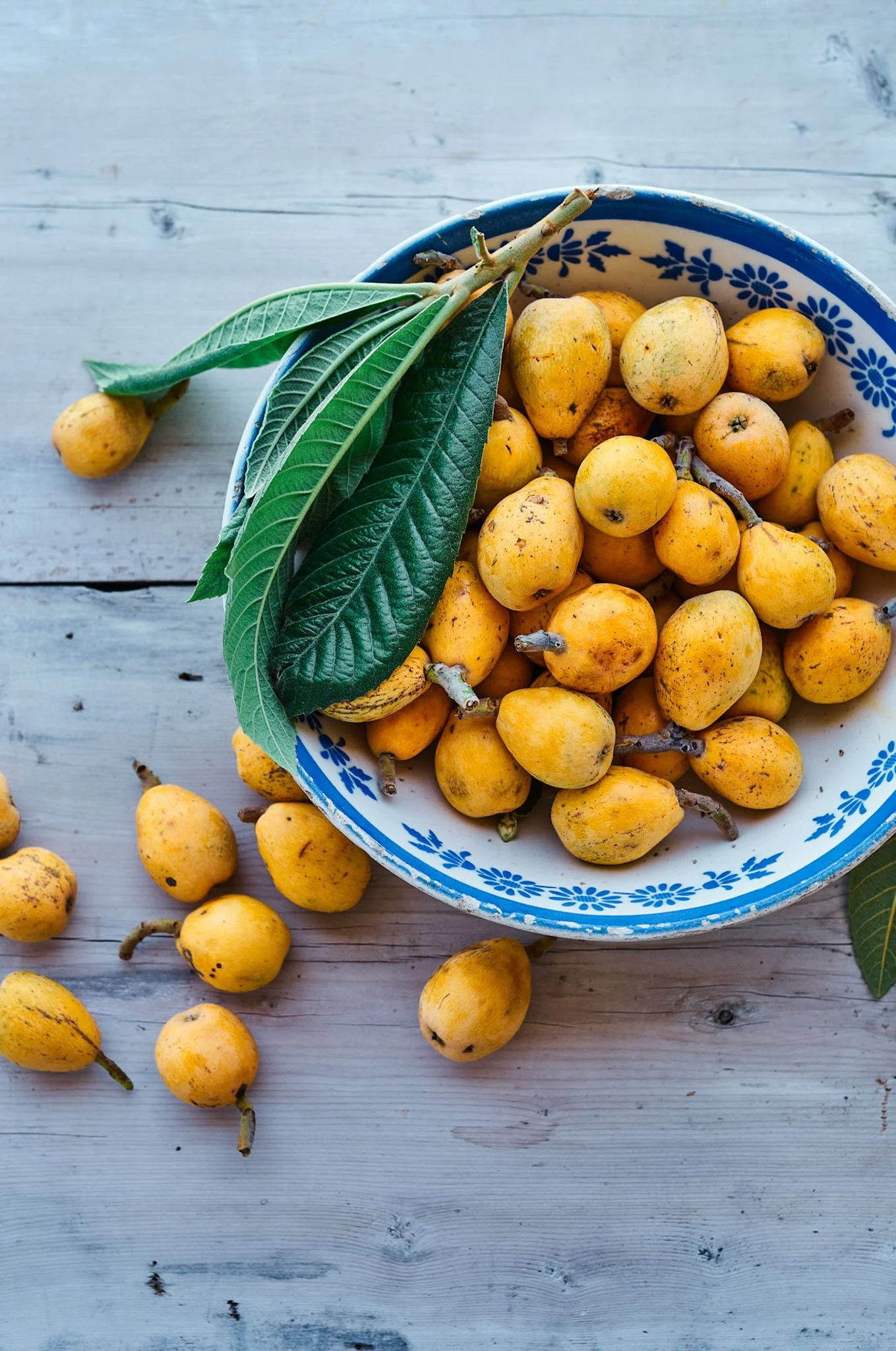Yellow loquats in a blue and white patterned bowl on a wooden surface, with some loquats and green leaves scattered around.