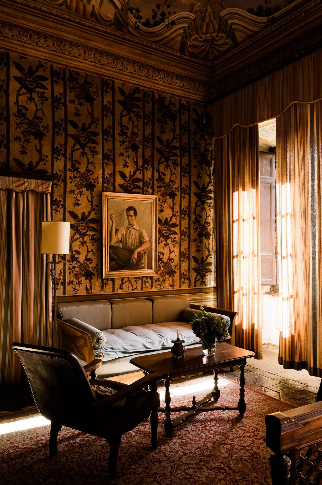 A vintage interior living room with ornate wallpaper, a beige sofa, a wooden coffee table with a flower vase, an upholstered armchair, a framed portrait on the wall, and curtains letting sunlight into the room.