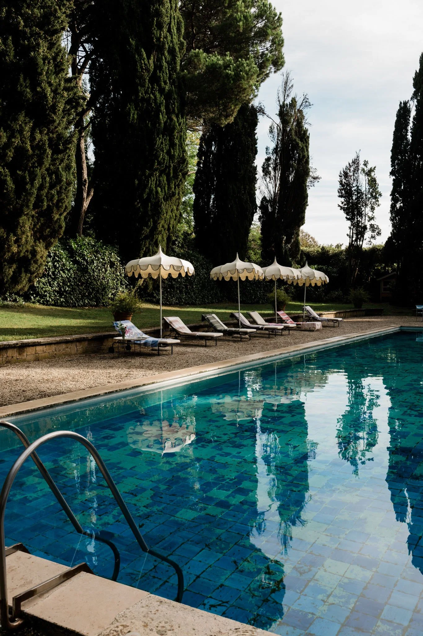 A swimming pool with blue tiles, surrounded by lounge chairs and white umbrellas, with tall trees and greenery in the background under a partly cloudy sky.
