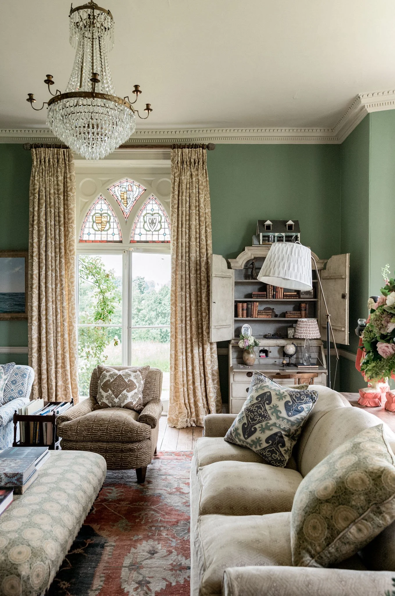 Living room with vintage furniture, patterned curtains, a stained glass window, a chandelier, bookshelves, and floral decorations.
