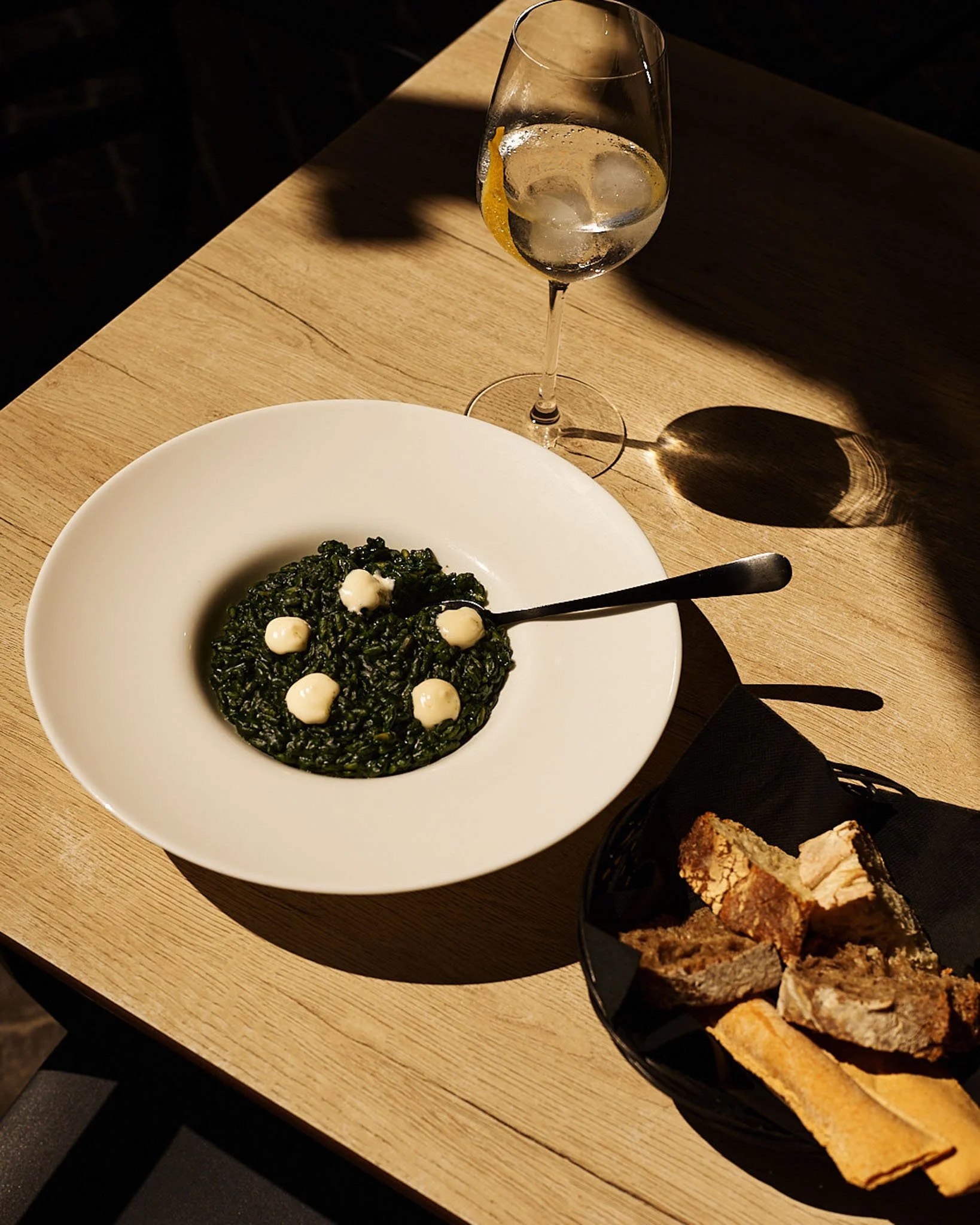 A bowl of black risotto with dollops of white sauce, a glass of sparkling water with lemon slices, and a basket of assorted bread slices on a wooden table.
