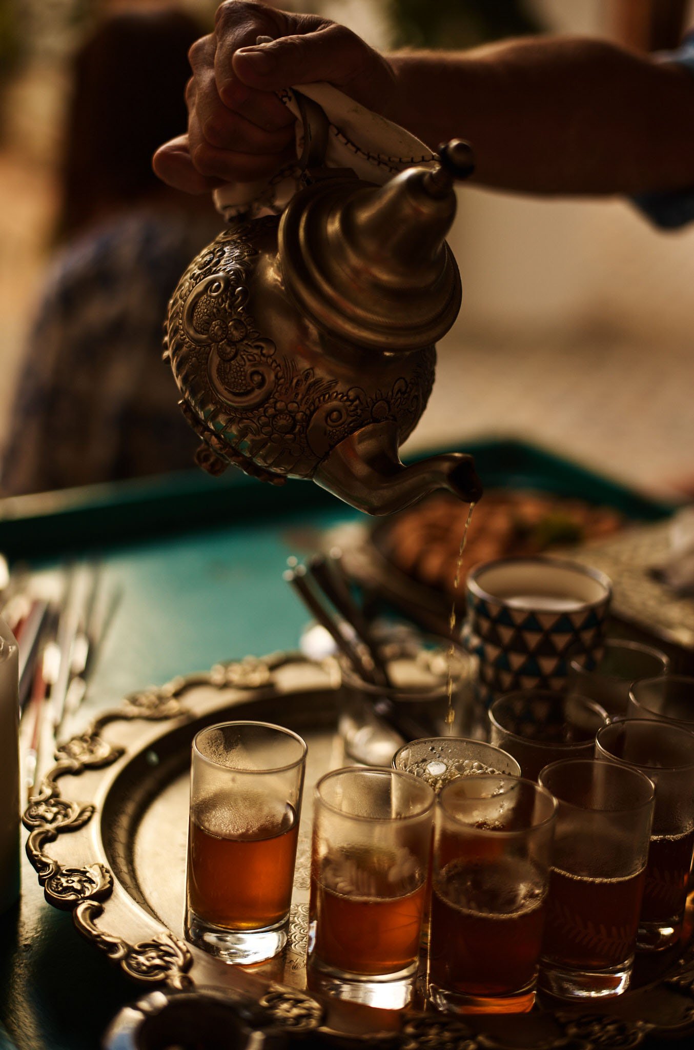 A person pouring tea from an ornate brass teapot into small glasses on a silver tray.