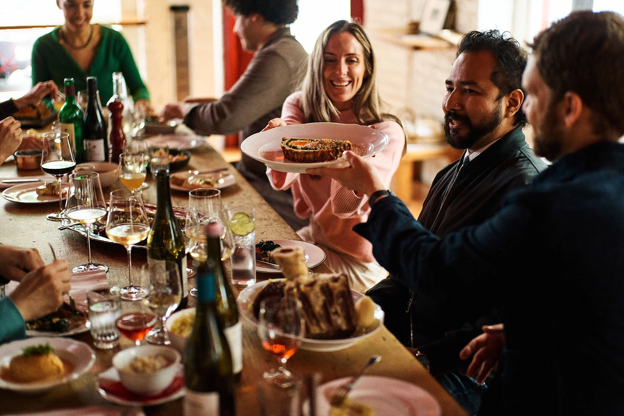 People gathered around a dinner table enjoying a meal, with a woman passing a dish to a man in the center.
