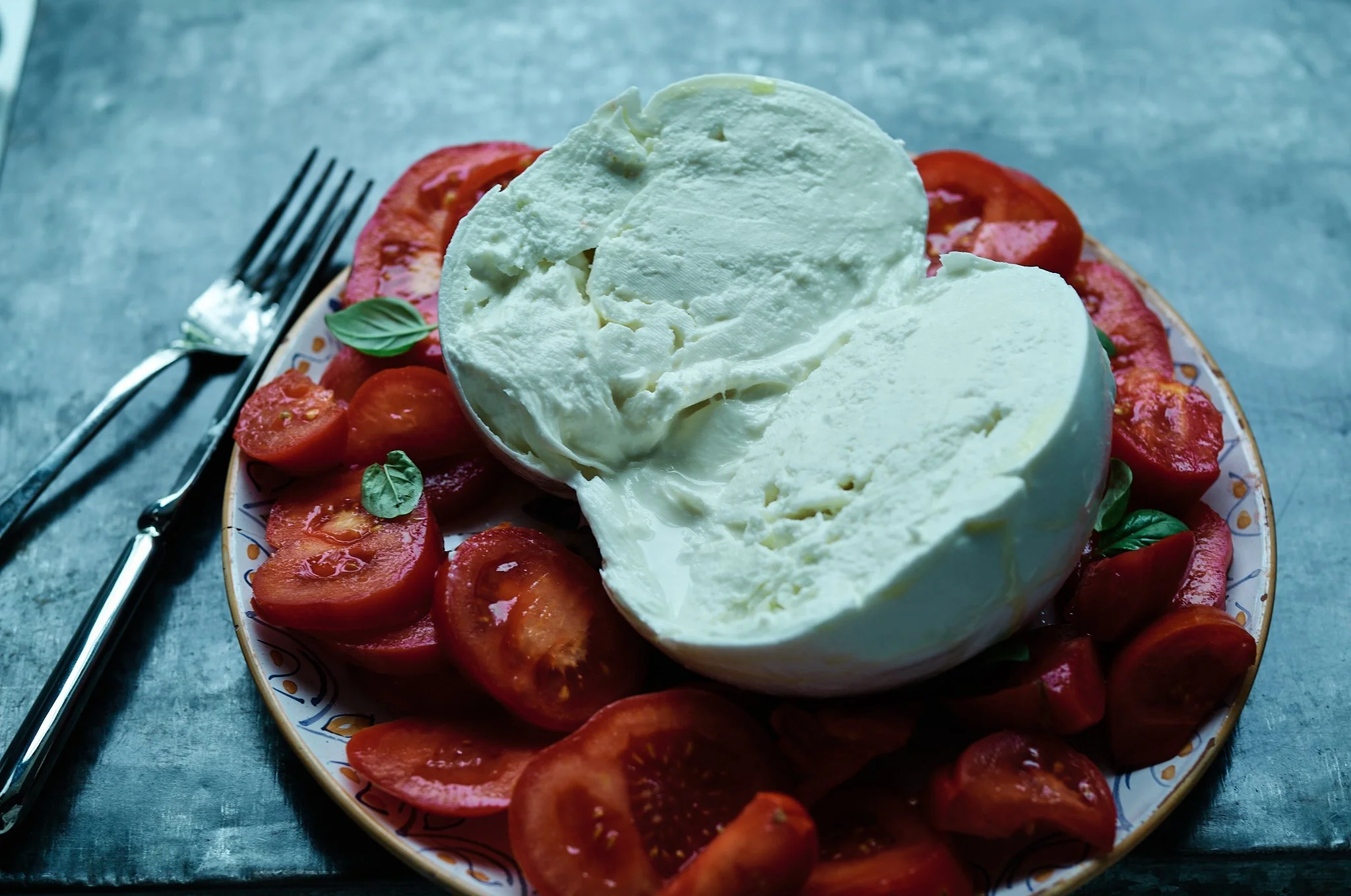 A bowl of fresh mozzarella cheese on a plate with sliced tomatoes and basil leaves, with a fork and knife beside the plate on a textured blue surface.