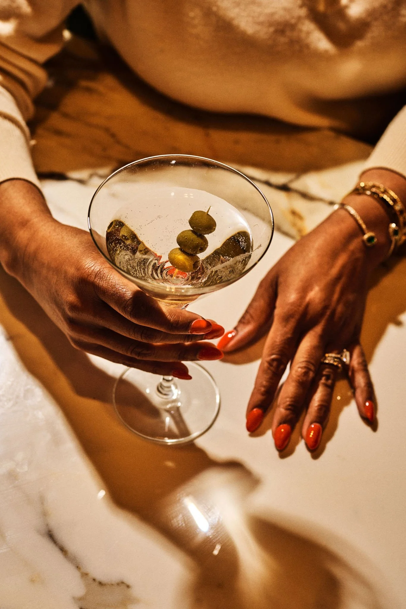 A person with orange nail polish and jewelry holds a cocktail glass with green olives, with a marble surface in the background.